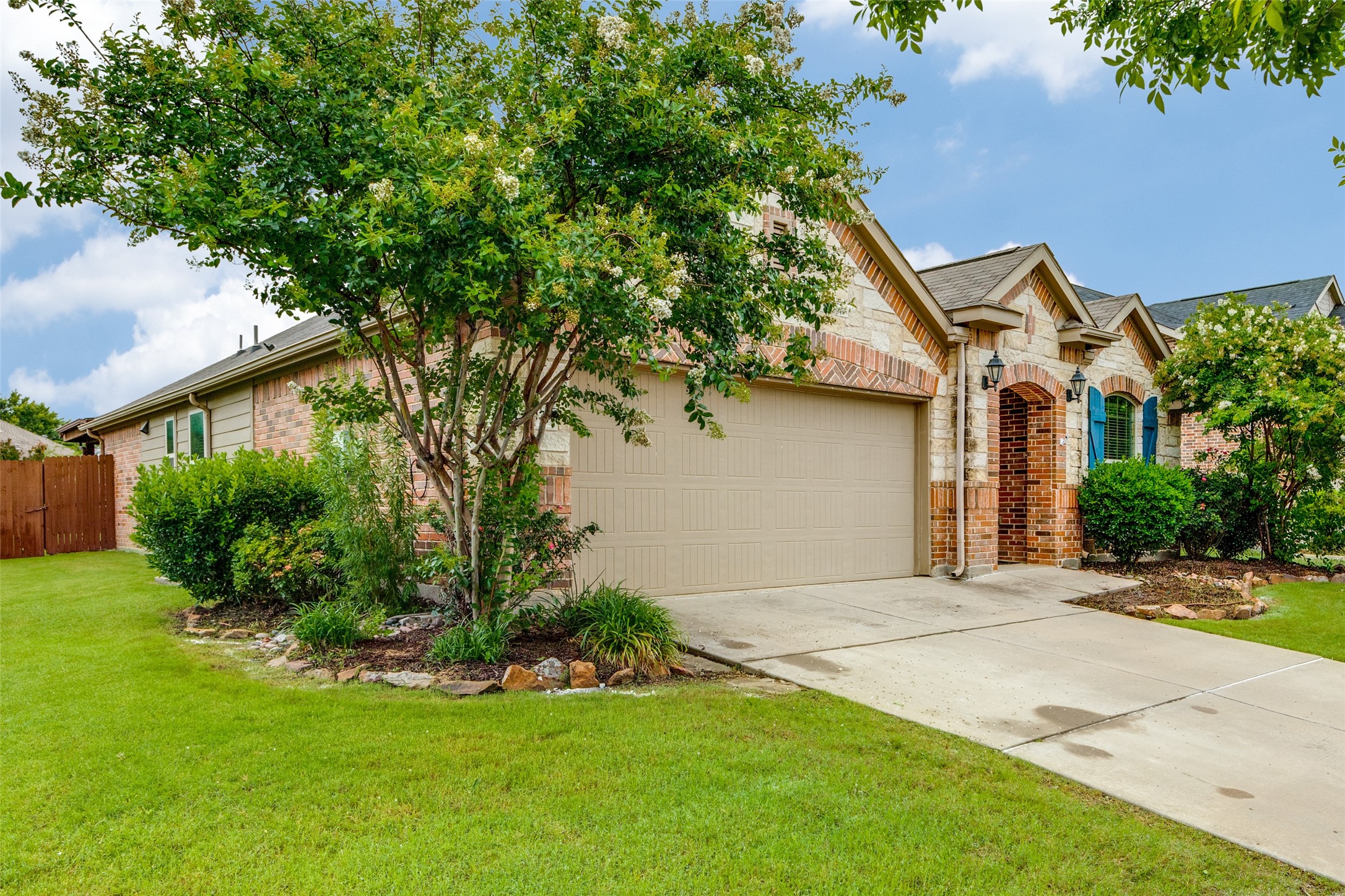 Brick-and-stone house with a two-car garage, blue shutters, and a flowering tree over the front lawn.