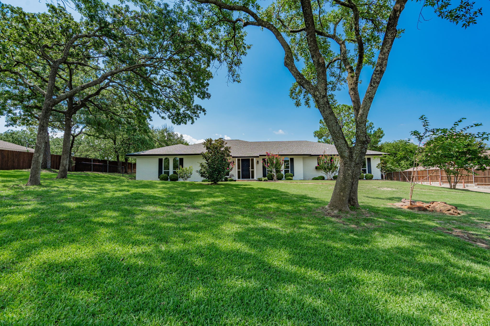 Single-story brick home framed by mature shade trees, with a wide lawn and fenced yard.