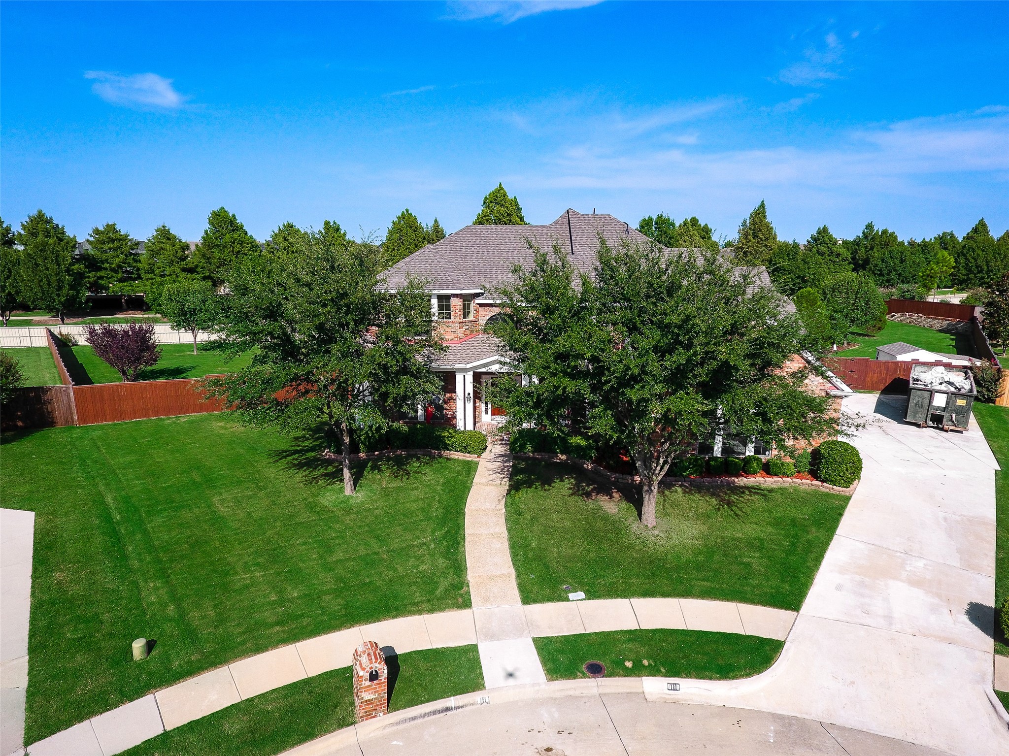Aerial view of a brick home with a broad green lawn, mature trees, and a curved sidewalk.
