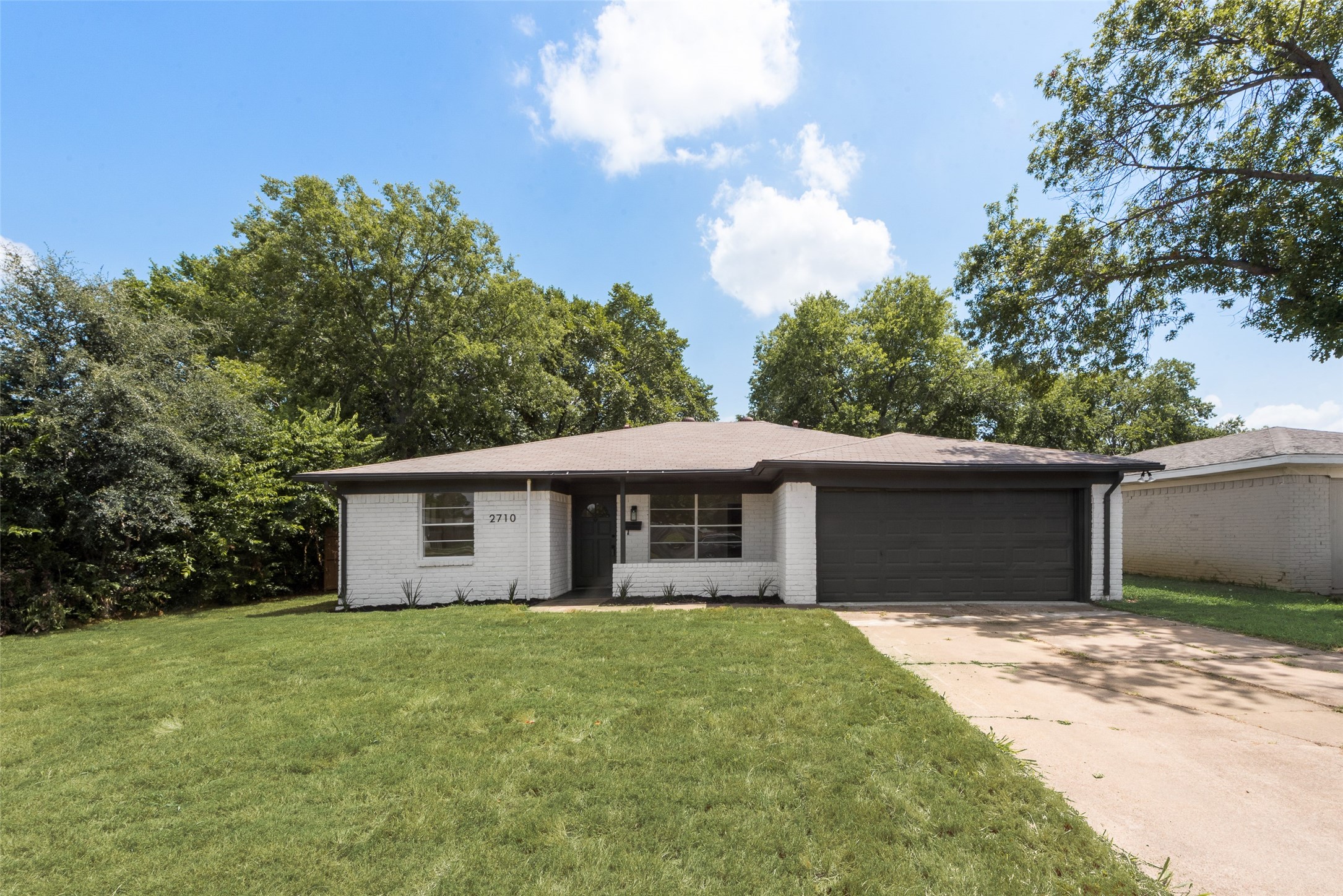 Single-story white brick house with a dark garage, broad lawn, and mature trees.
