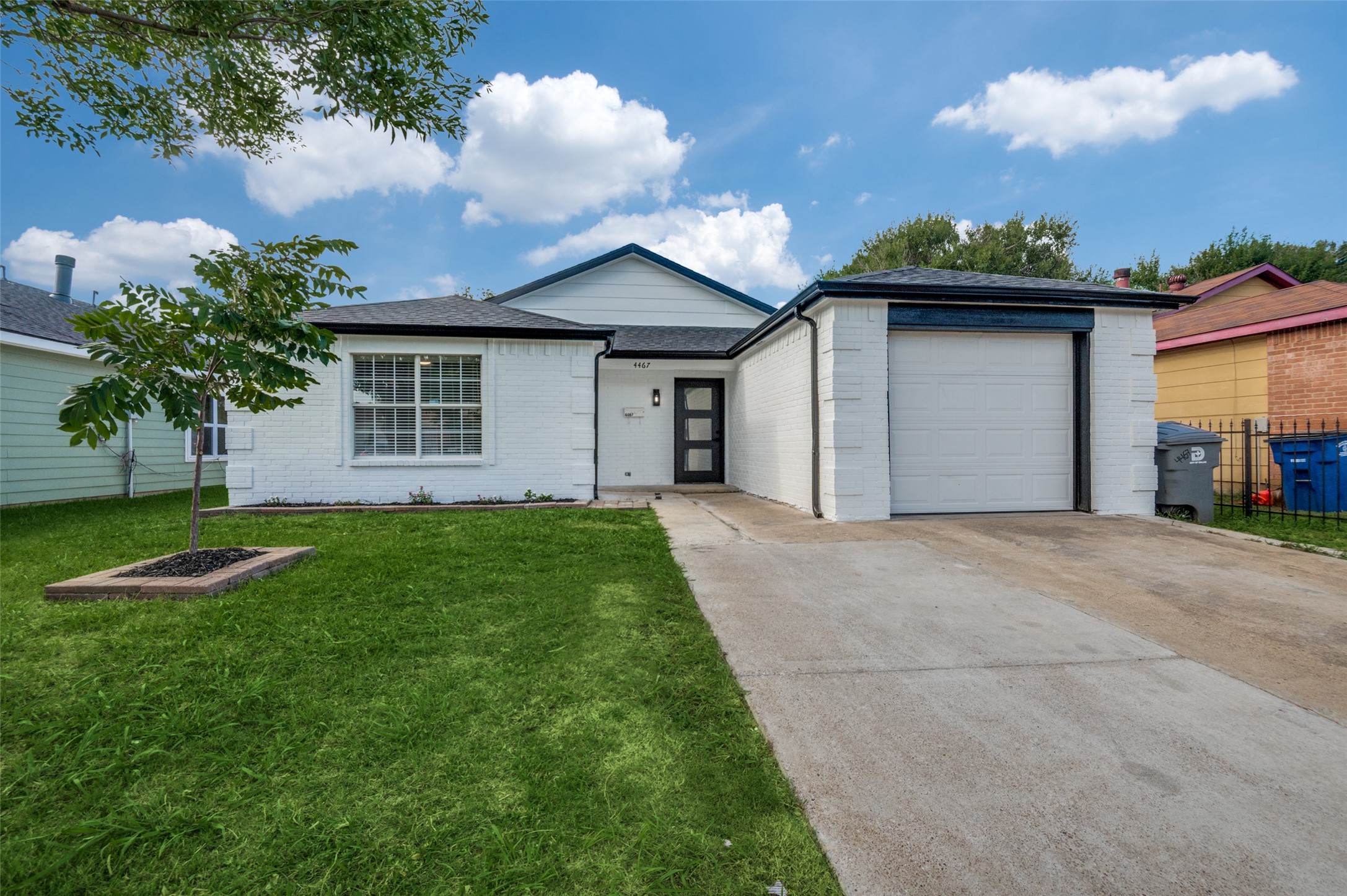 White brick single-story house with an attached garage, broad driveway, and a young tree in the front lawn.