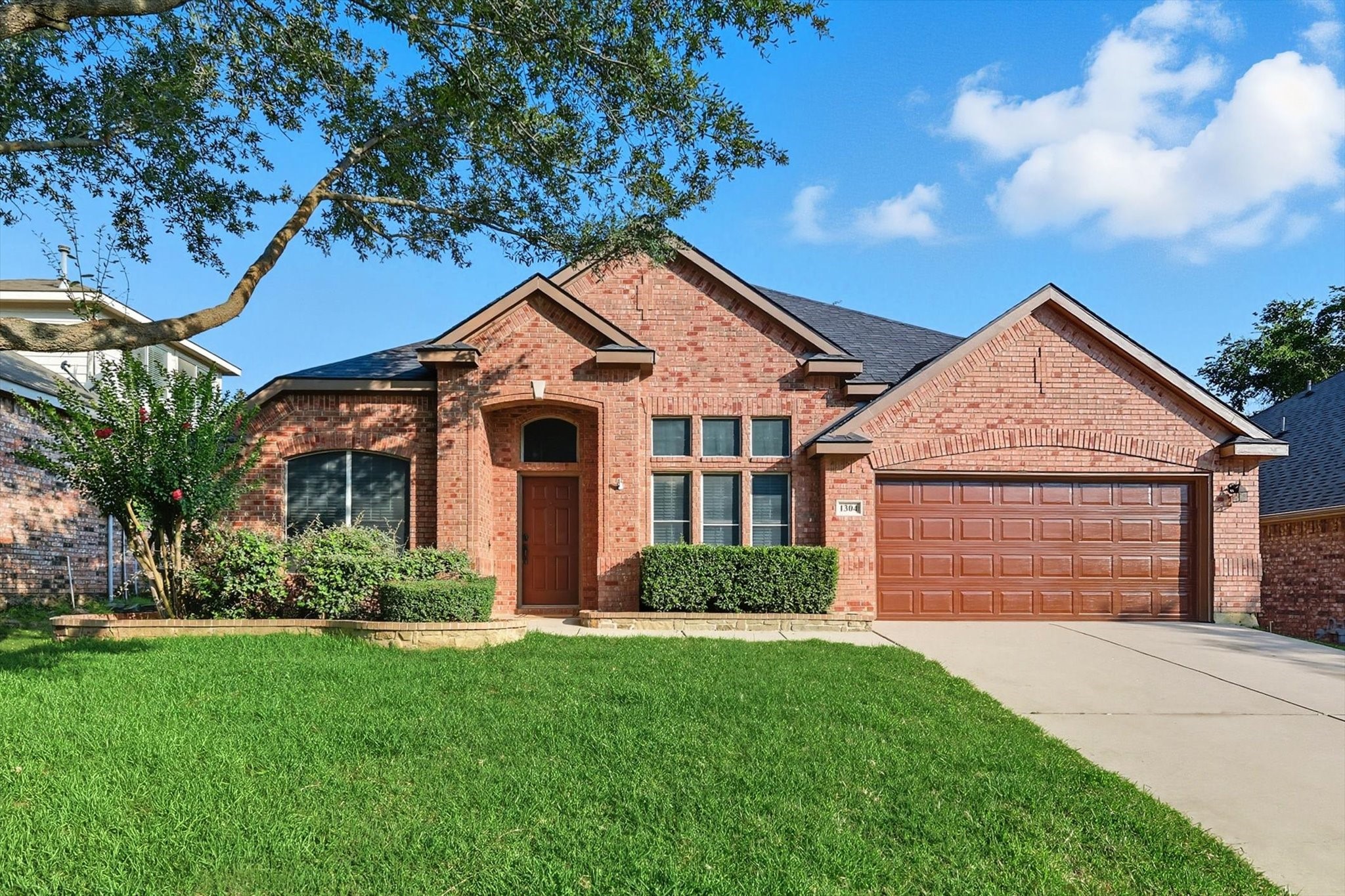 Red-brick house with a two-car garage, broad green lawn, and concrete driveway.