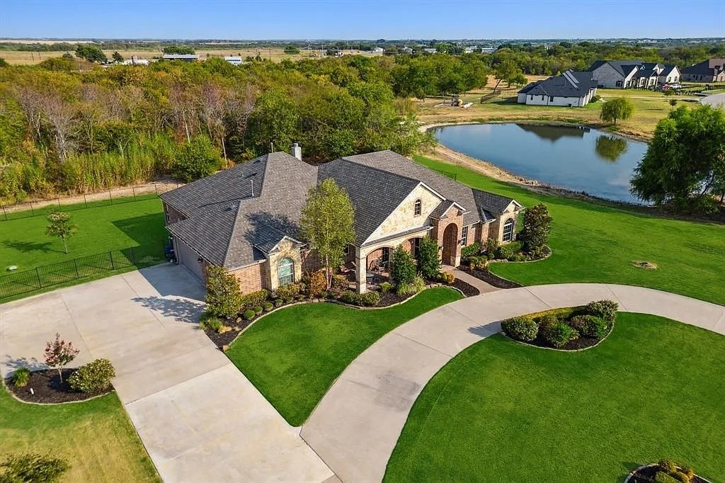 Aerial view of a brick-and-stone house with a curved driveway beside a pond.