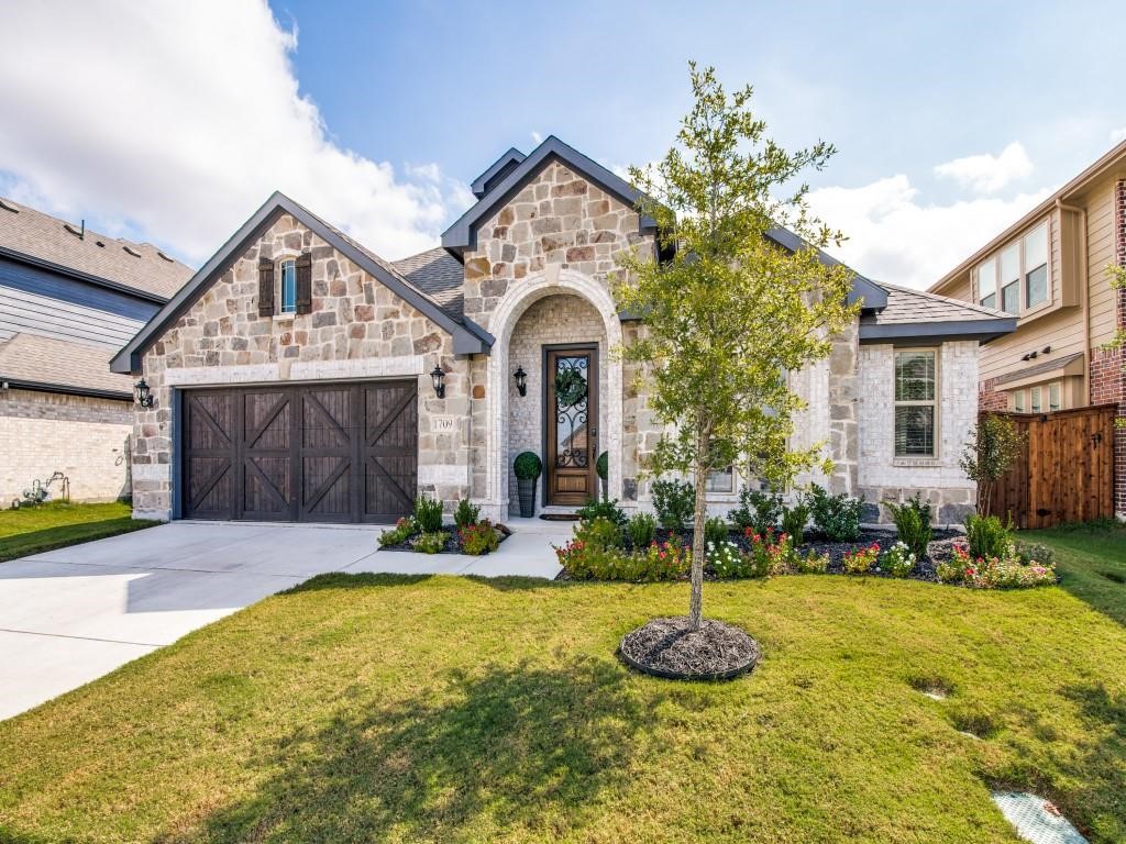 Stone-front house with an arched entry, dark double garage door, and landscaped front lawn.