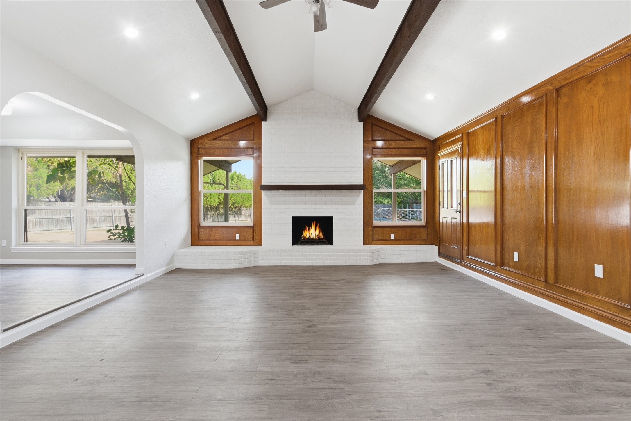 Vacant living room with vaulted beam ceiling, white brick fireplace, and warm wood-paneled wall.