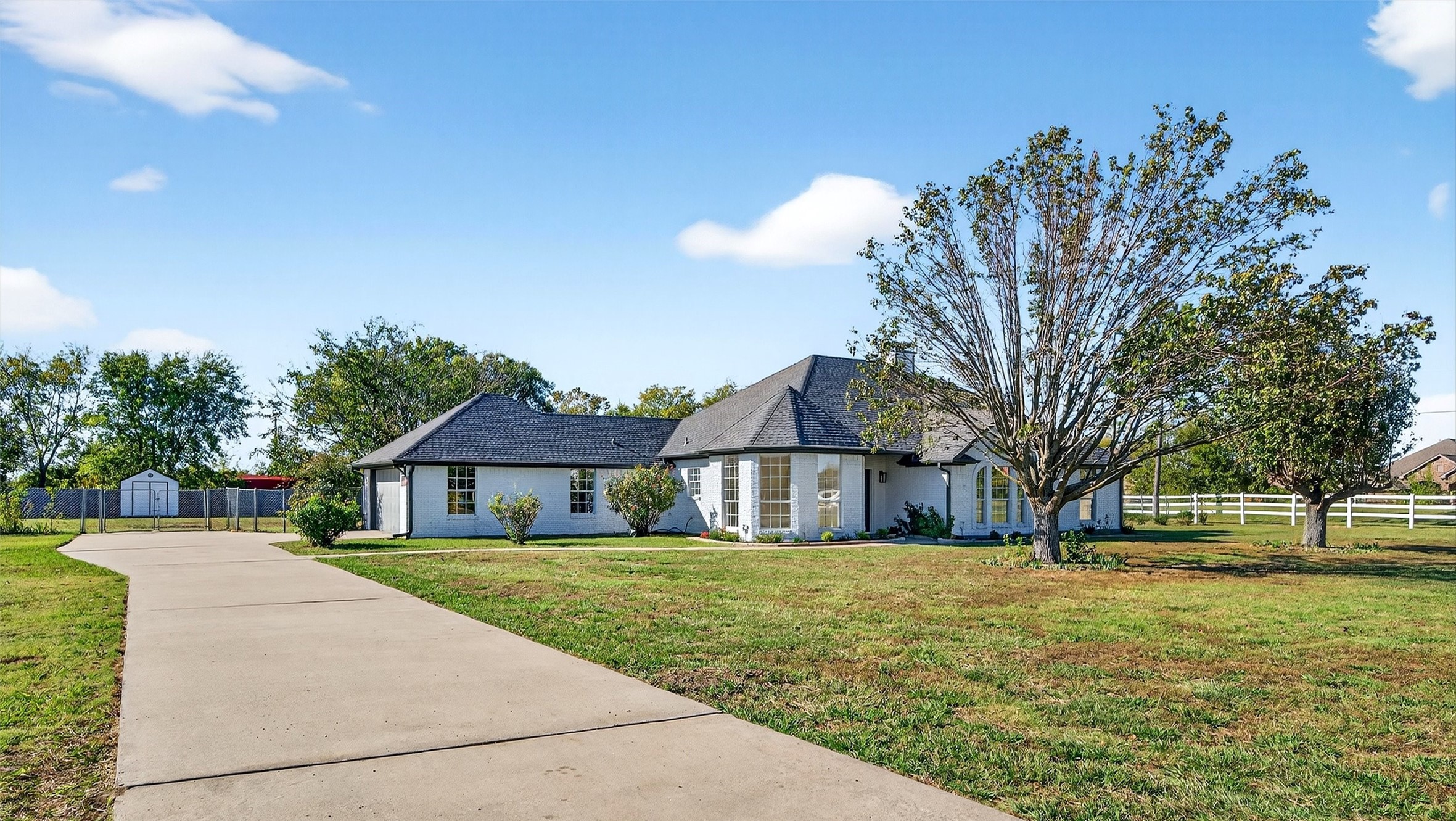 White brick single-story home with a curved driveway, mature trees, and white fencing.