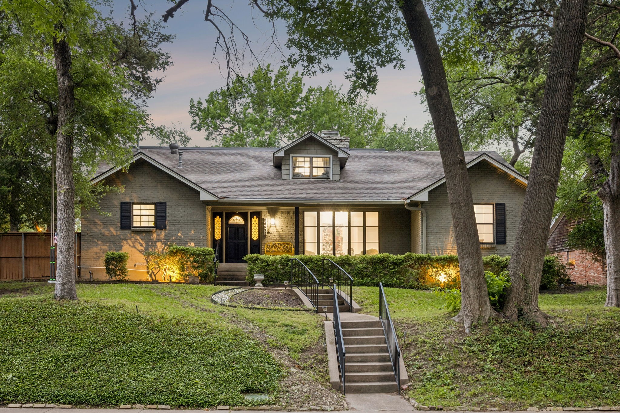 Gray brick house with a dormer, warmly lit windows, and a long staircase through mature trees.