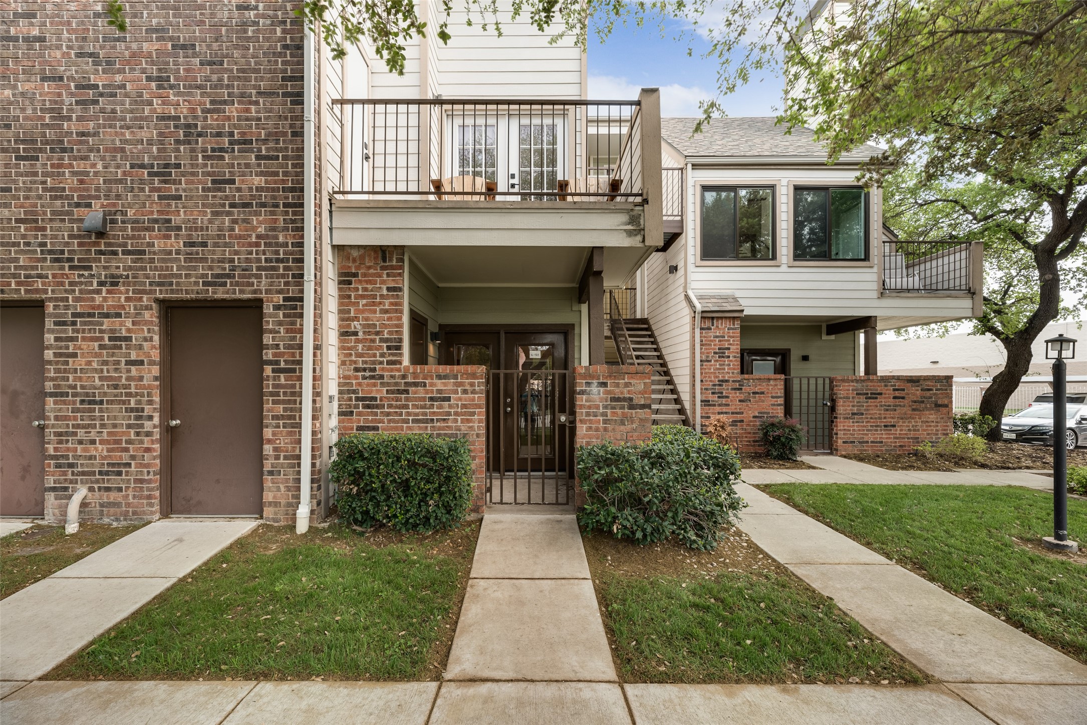 Multi-unit building entrance with brick walls, raised balconies, exterior stairs, and landscaped walkways.