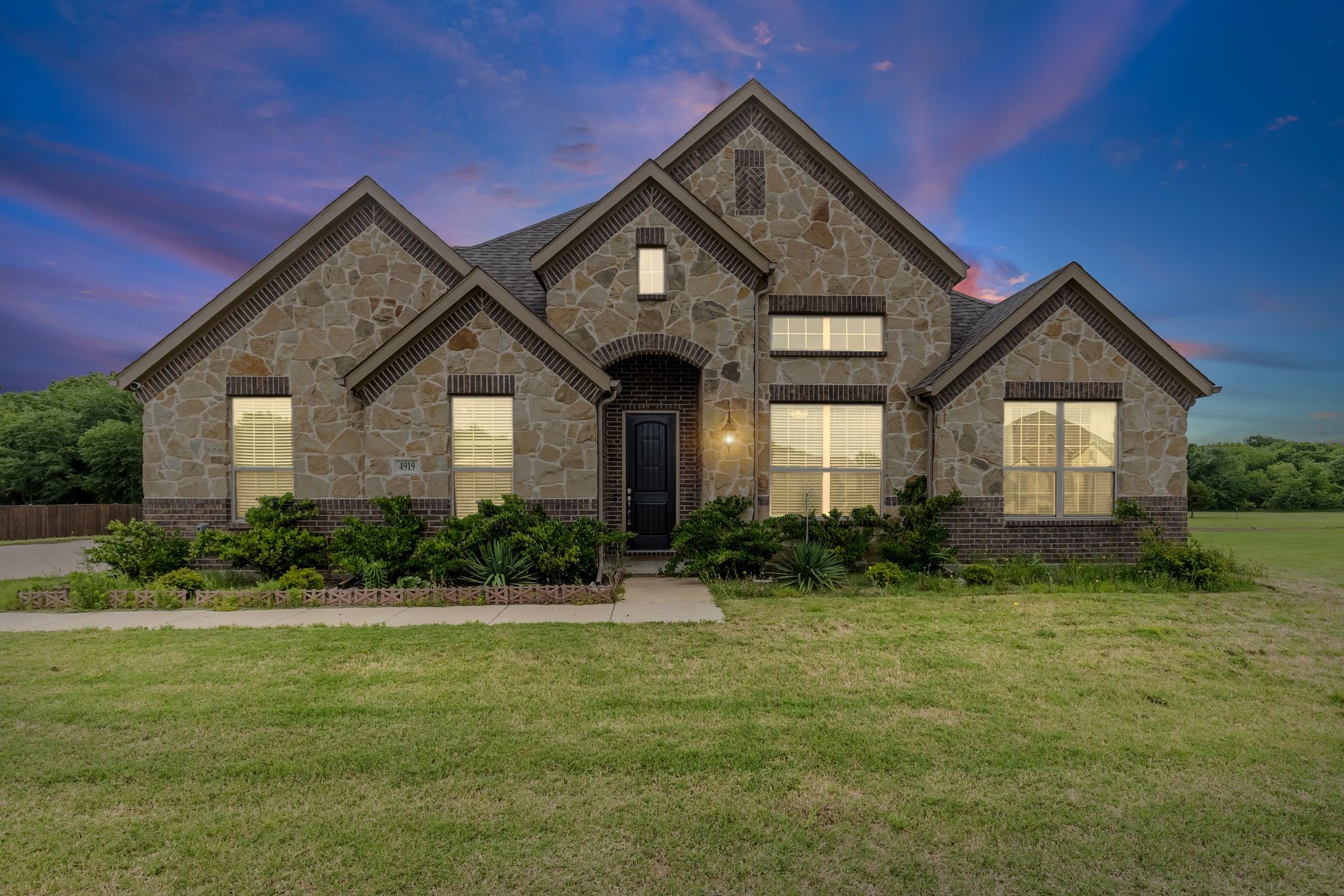 Two-story stone house with multiple gables, illuminated windows, landscaped shrubs, and a broad lawn at dusk.