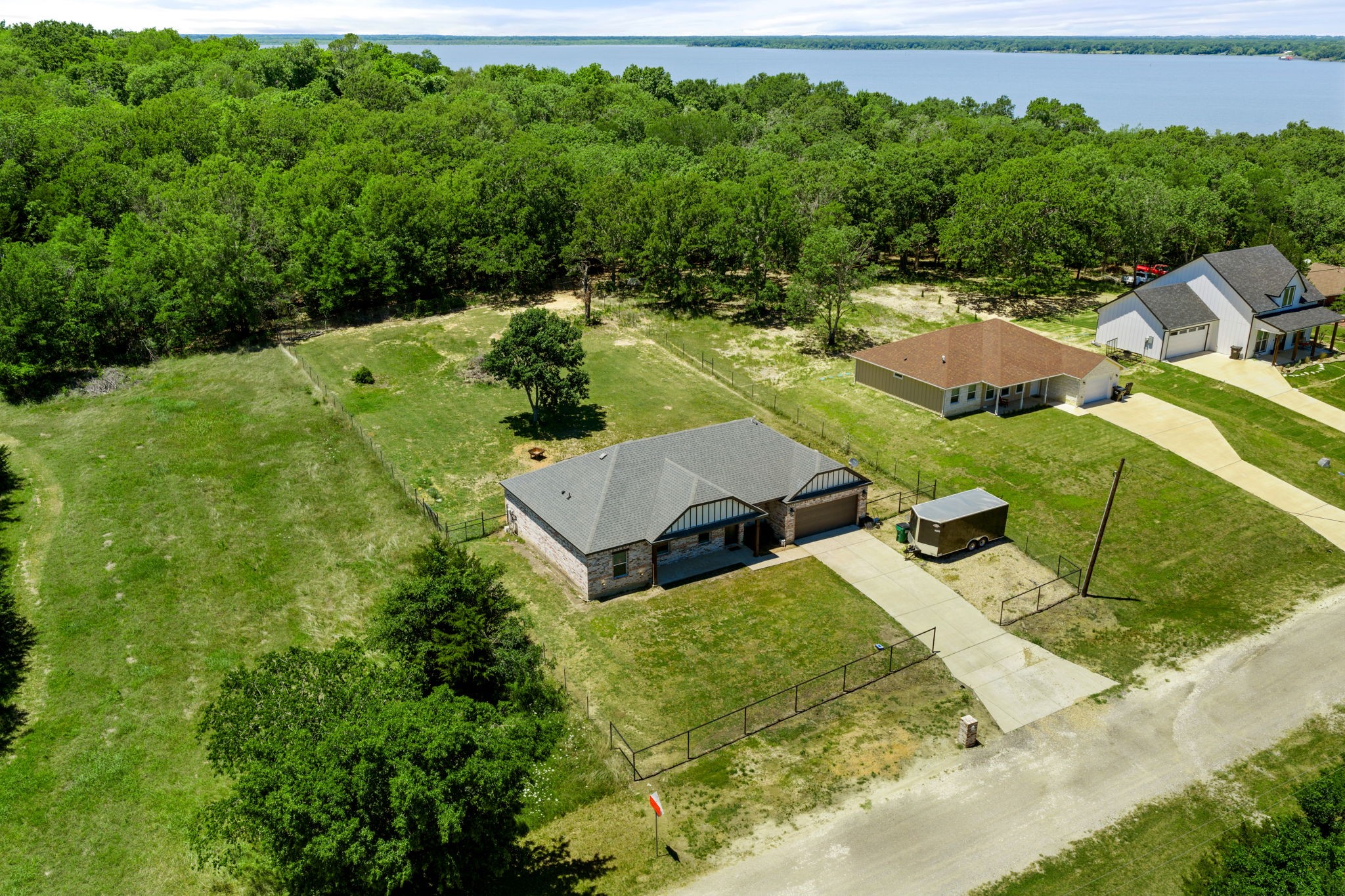 Aerial view of a brick home with a fenced grassy lot, neighboring houses, dense trees, and a lake beyond.