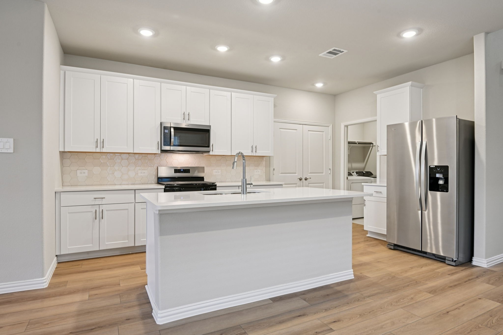 Modern white kitchen with a large island, stainless steel refrigerator, and wood flooring.