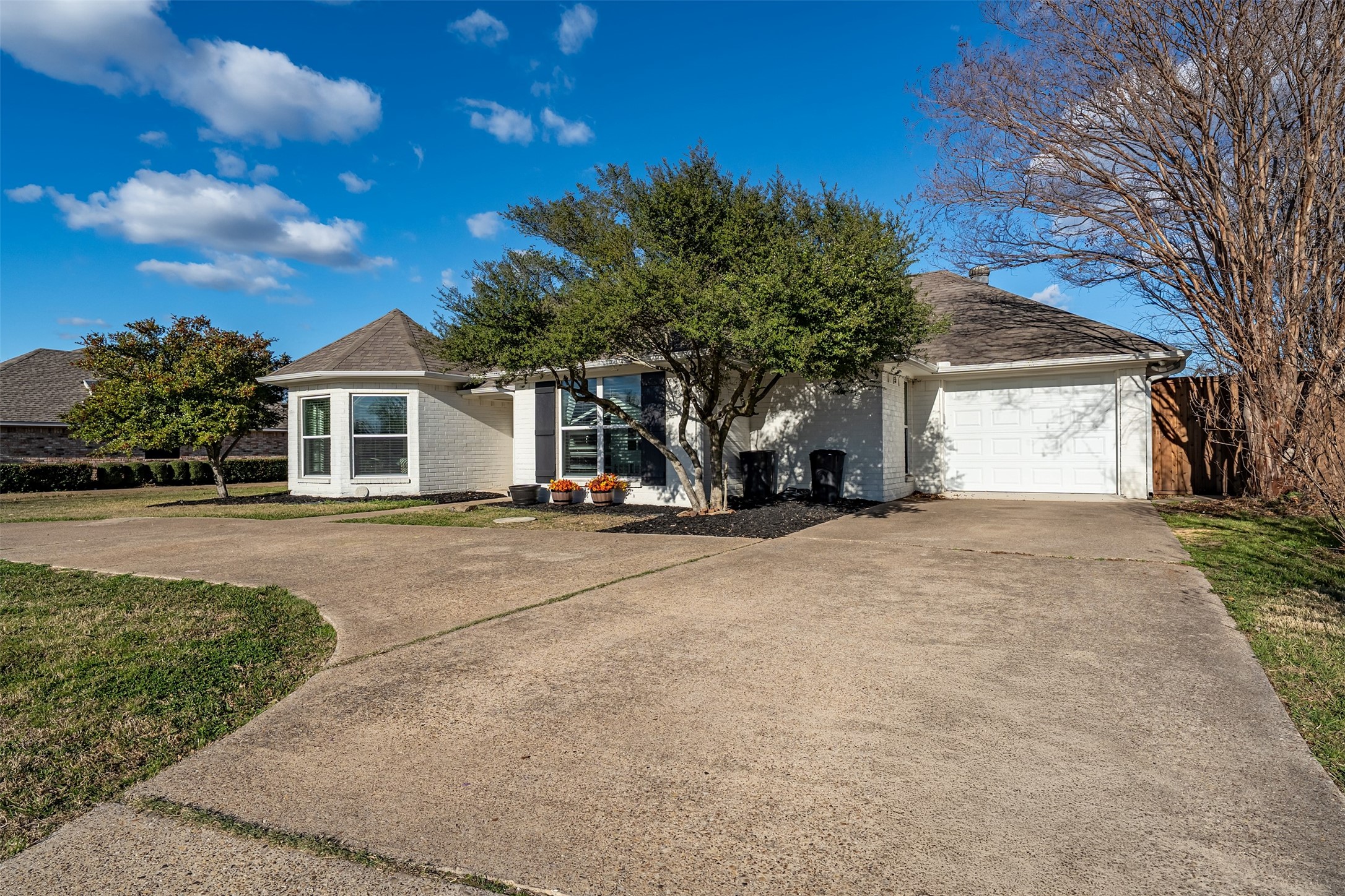 Light brick house with an attached garage, broad driveway, and mature trees in the front yard.