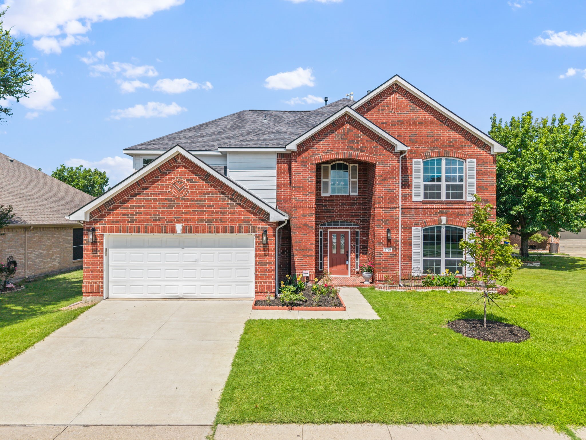 Two-story red brick house with a white double garage, arched windows, and a landscaped lawn.