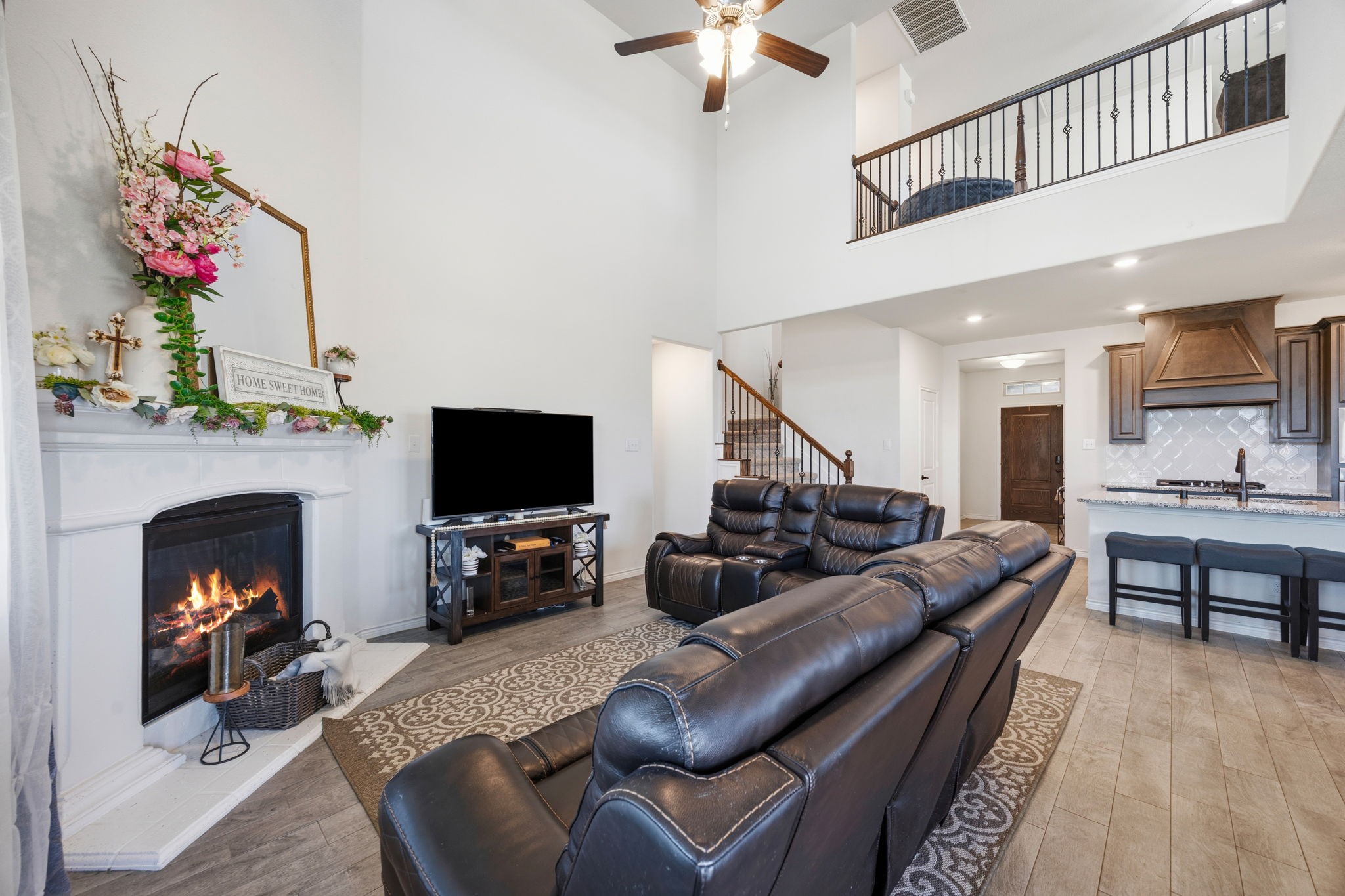 Open-plan living room with a leather sectional, lit fireplace, and tall ceiling overlooking the kitchen.
