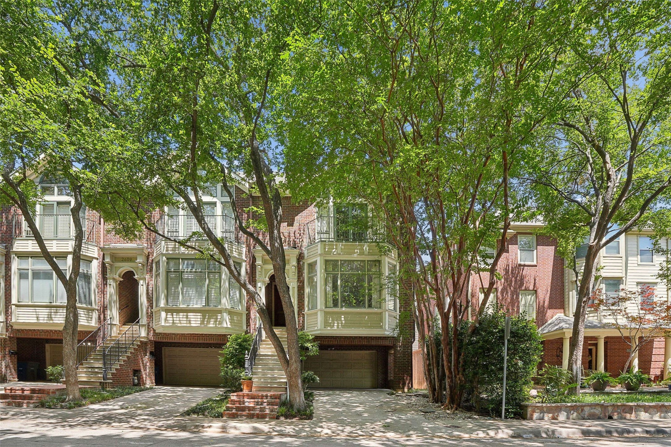 Brick townhomes with bay windows and garages, shaded by mature leafy trees.