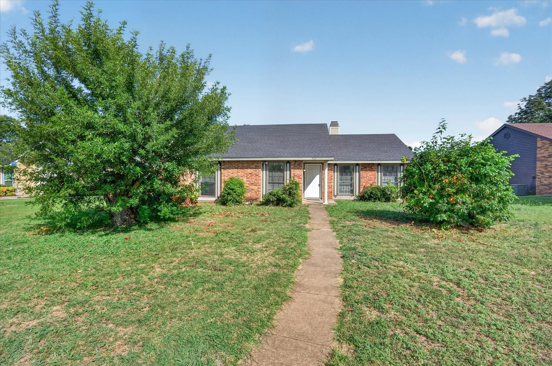 Single-story brick house with a white front door, concrete walkway, and mature trees in the yard.