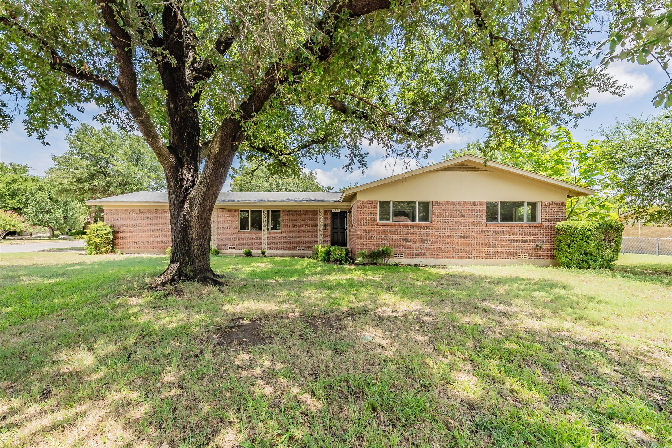 Single-story brick house behind a lawn, shaded by a large mature tree.
