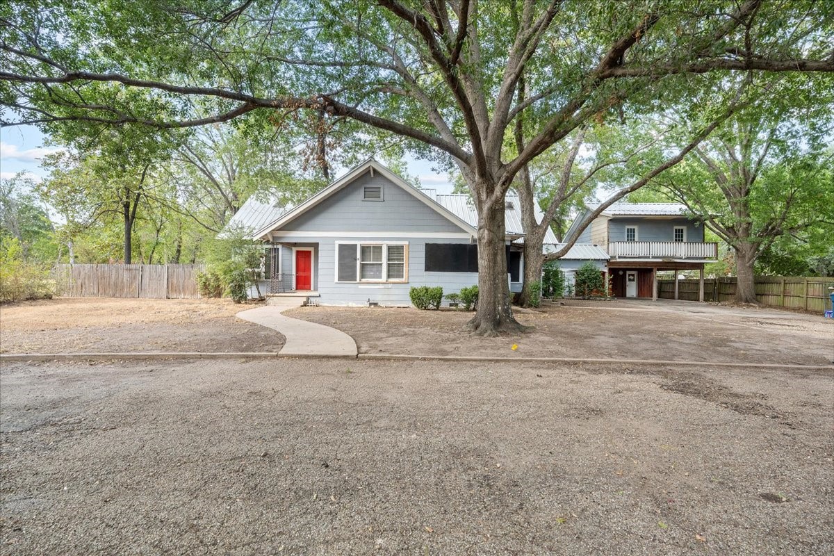Gray house with a bright red front door beneath a large shade tree, with a raised porch to the right.