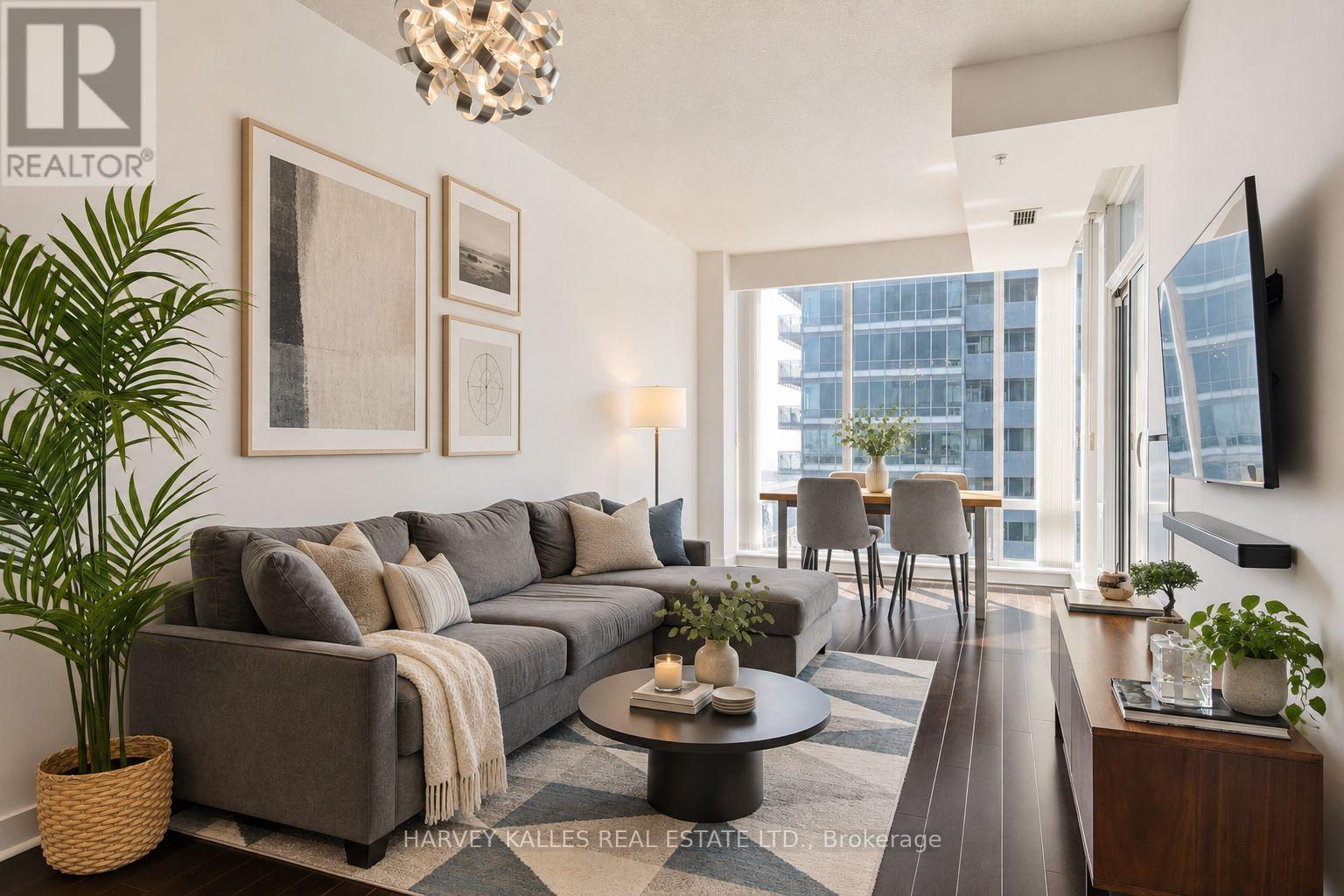 Staged condo living room with a gray sectional, dining nook, and large windows overlooking office buildings.