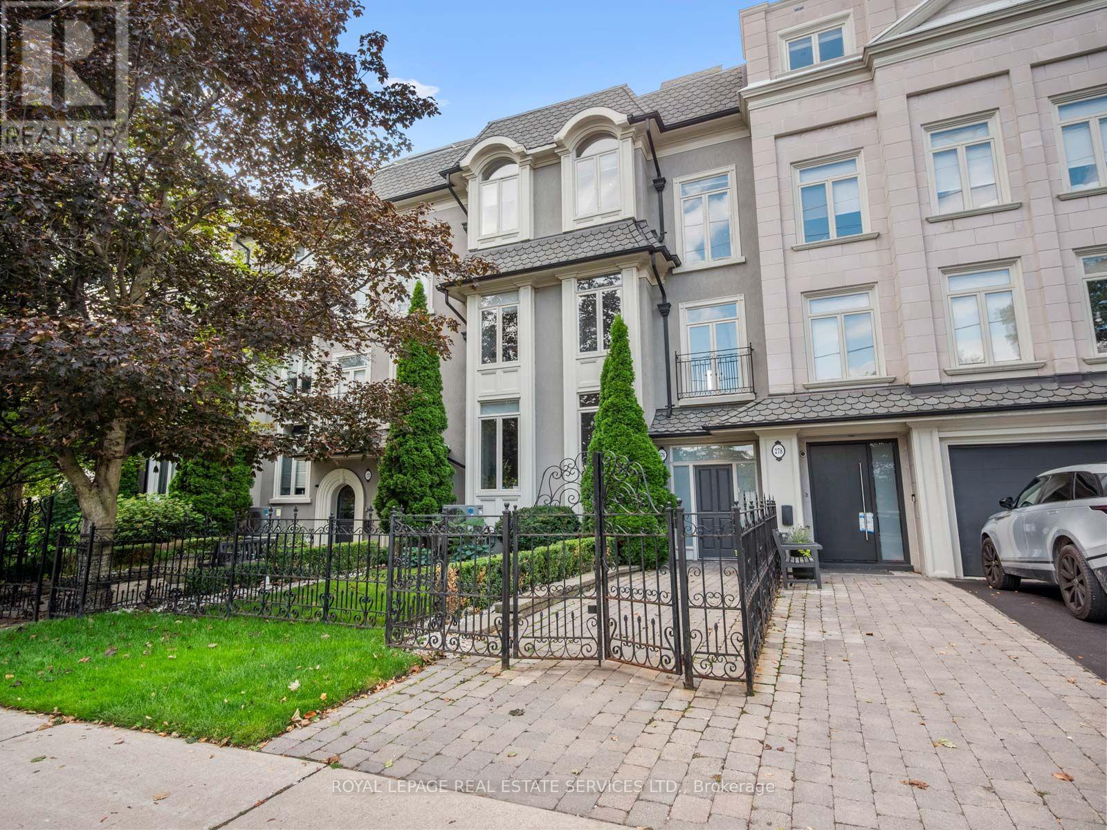 Attached multi-storey homes with ornate rooflines, fenced gardens, and a paved driveway beneath autumn-colored trees.