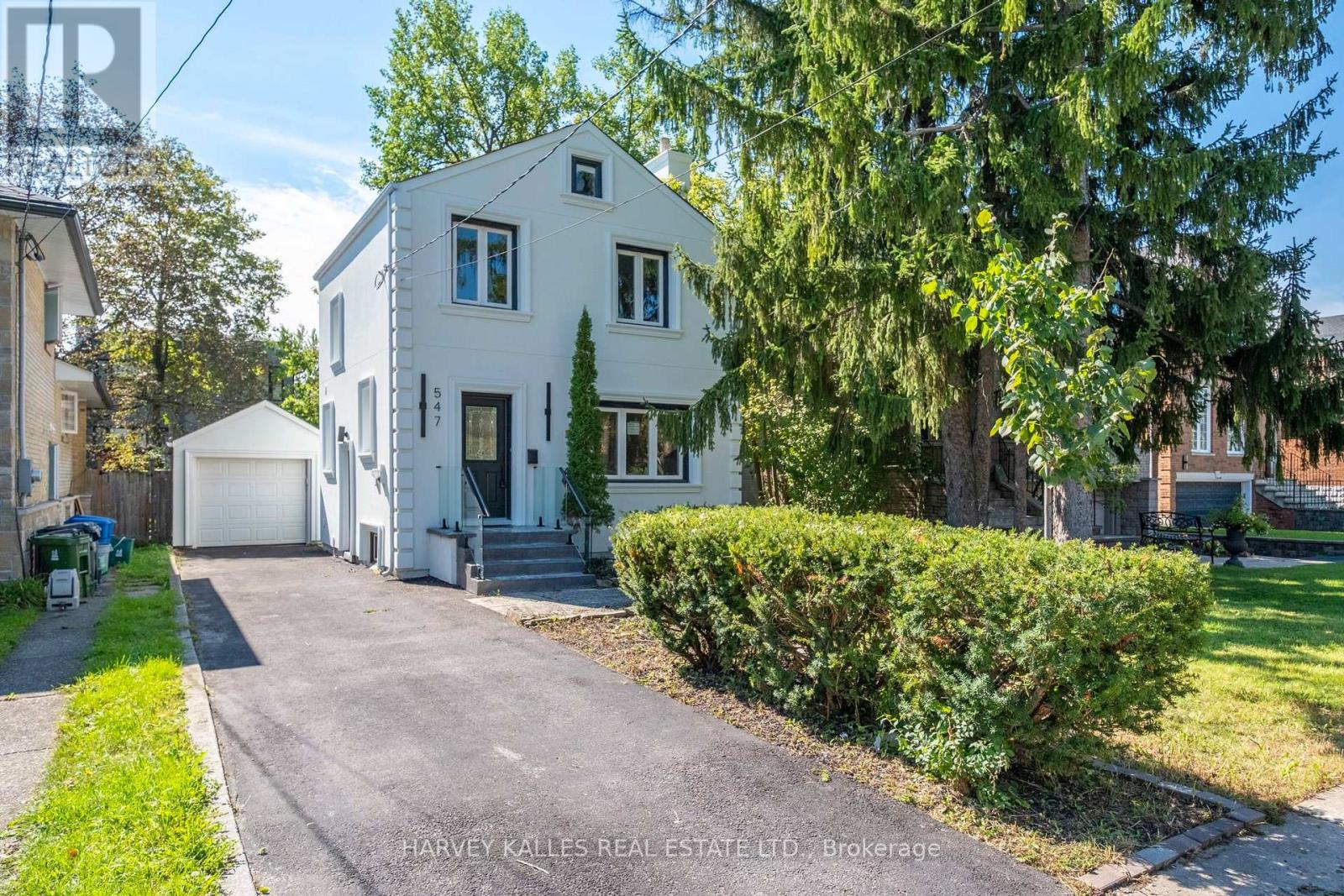 White two-story house with a detached garage, paved driveway, and mature trees.