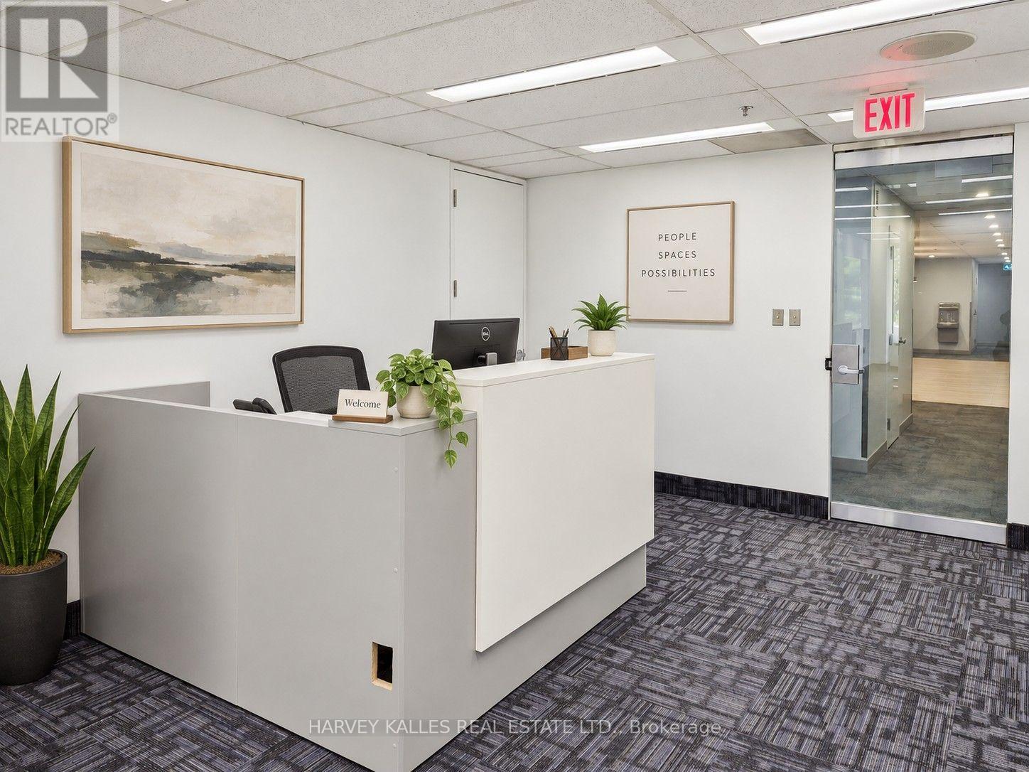 Office reception with a white front desk, leafy plants, framed artwork, and glass exit door.