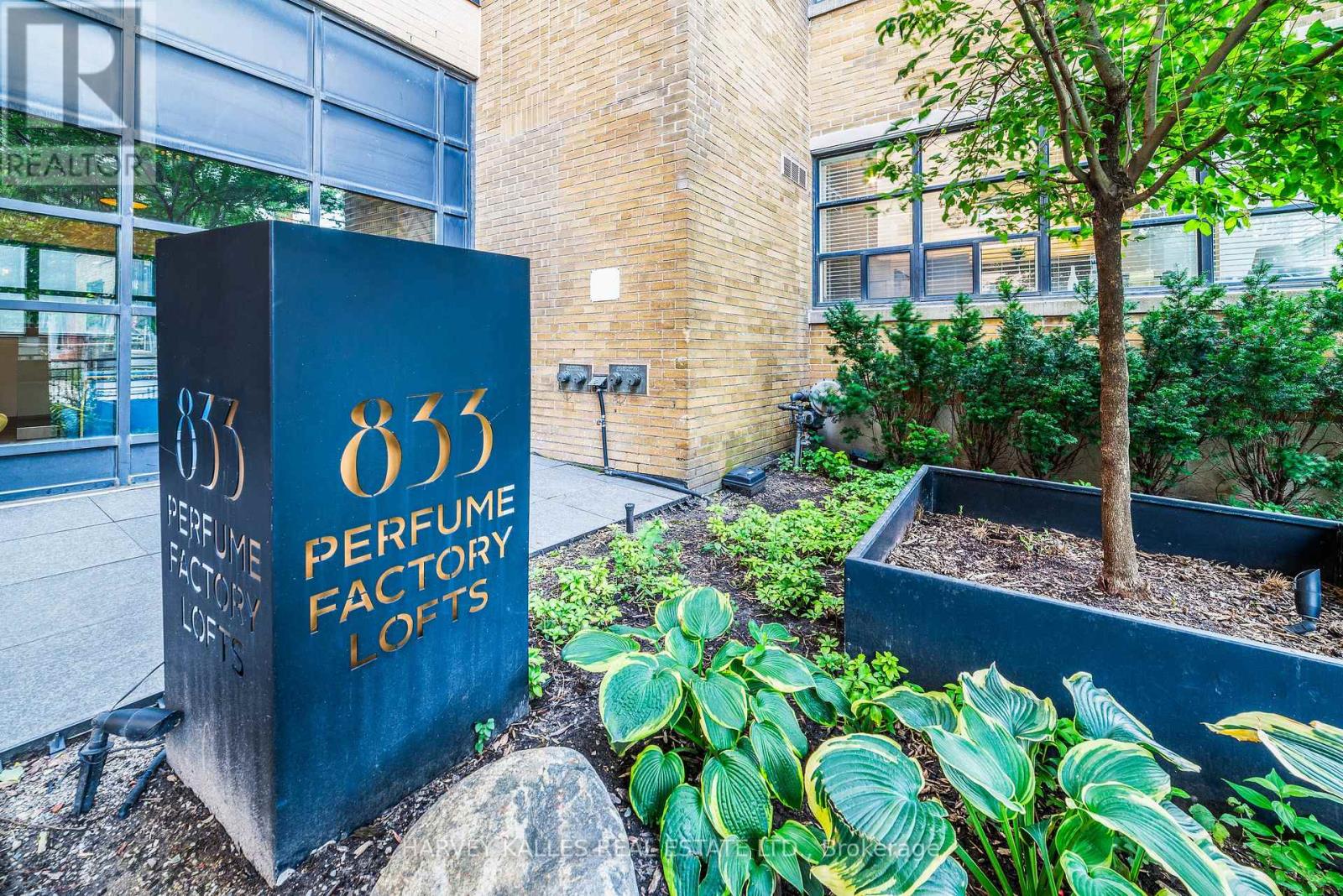 Perfume Factory Lofts entrance with an 833 sign, brick walls, blue-framed windows, and landscaped planting beds.