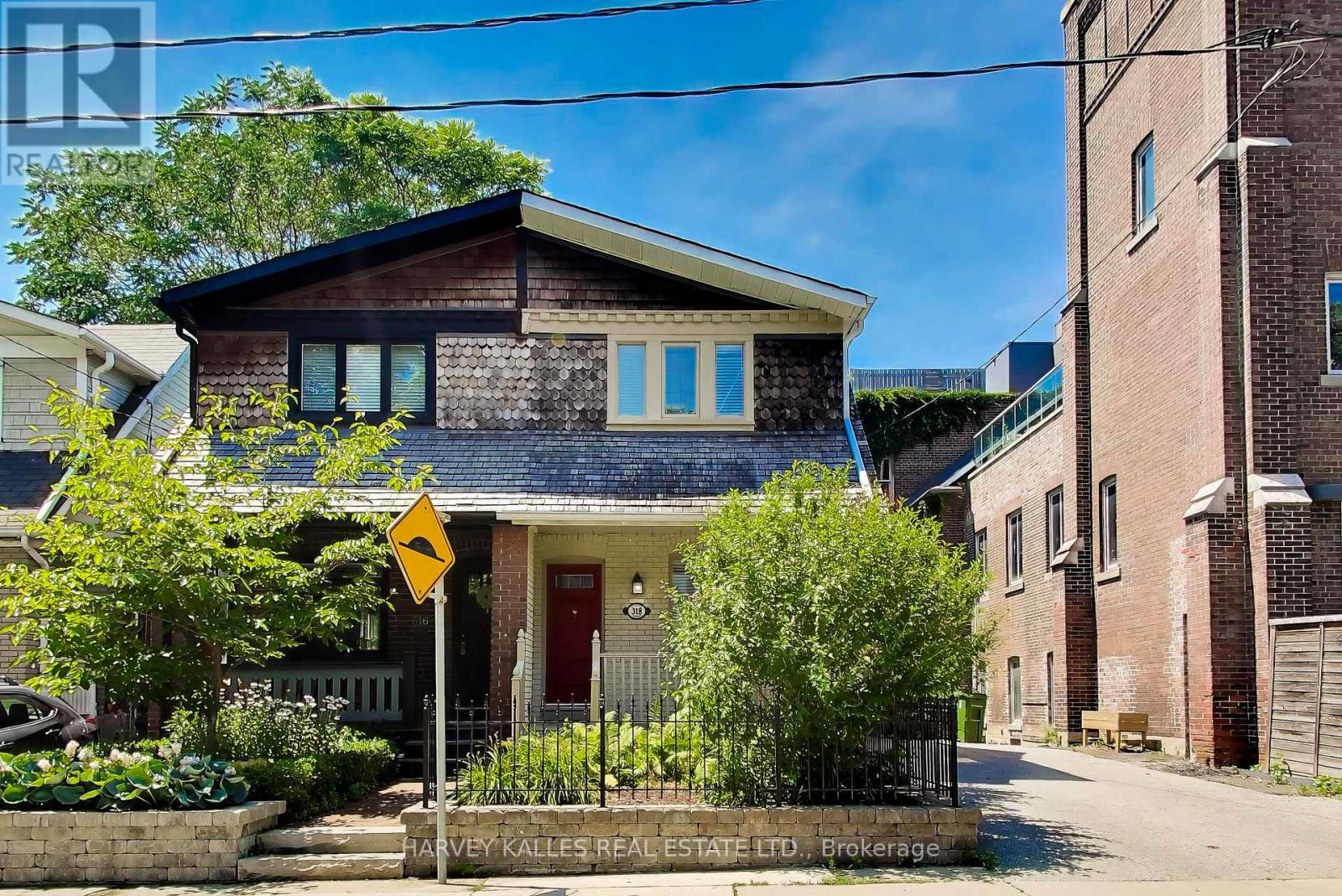 Dark-shingled two-story house with a red door, leafy front garden, and black metal fence.