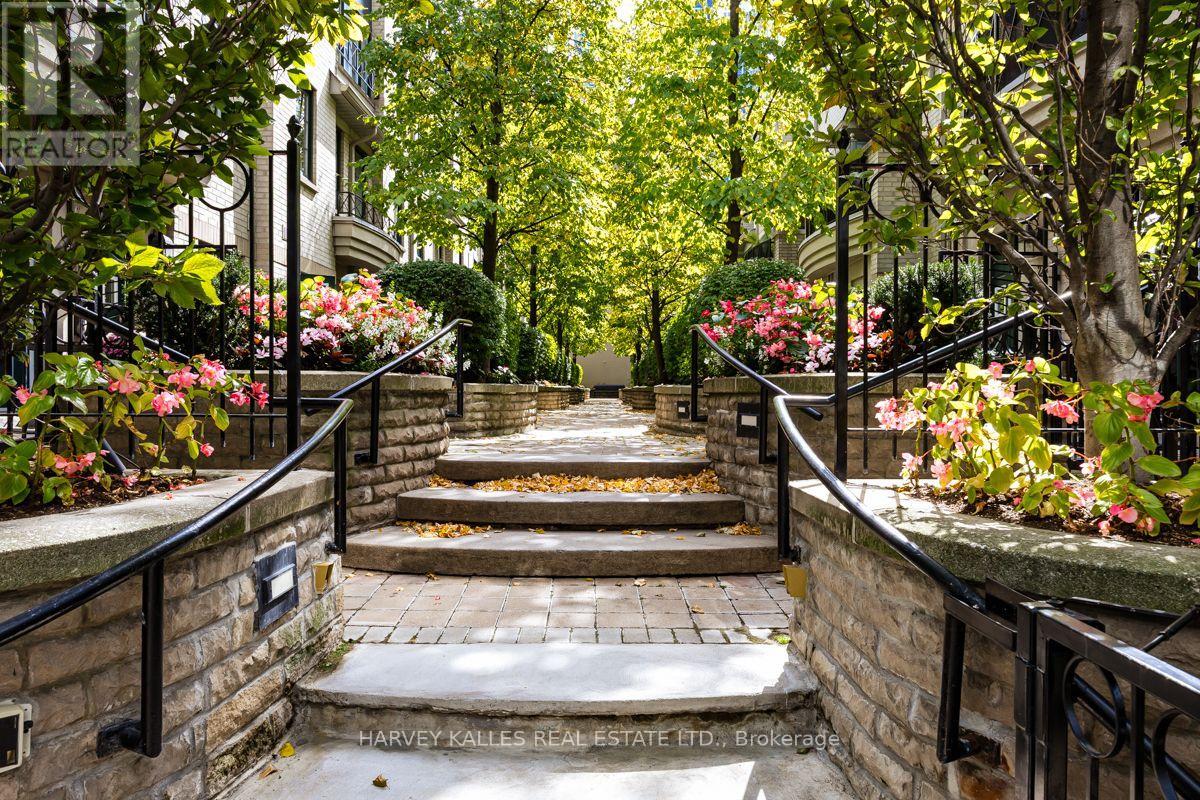 Residential building entrance reached by a stone walkway, steps, black railings, flowering shrubs, and mature trees.