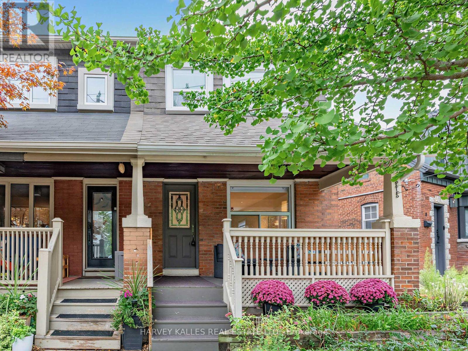 Brick house with a covered porch, white railings, and pink flowers beneath leafy branches.