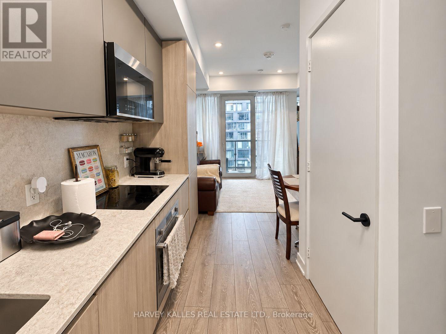 Modern condo kitchen with light wood cabinets and stone counters, opening toward a dining area and balcony.