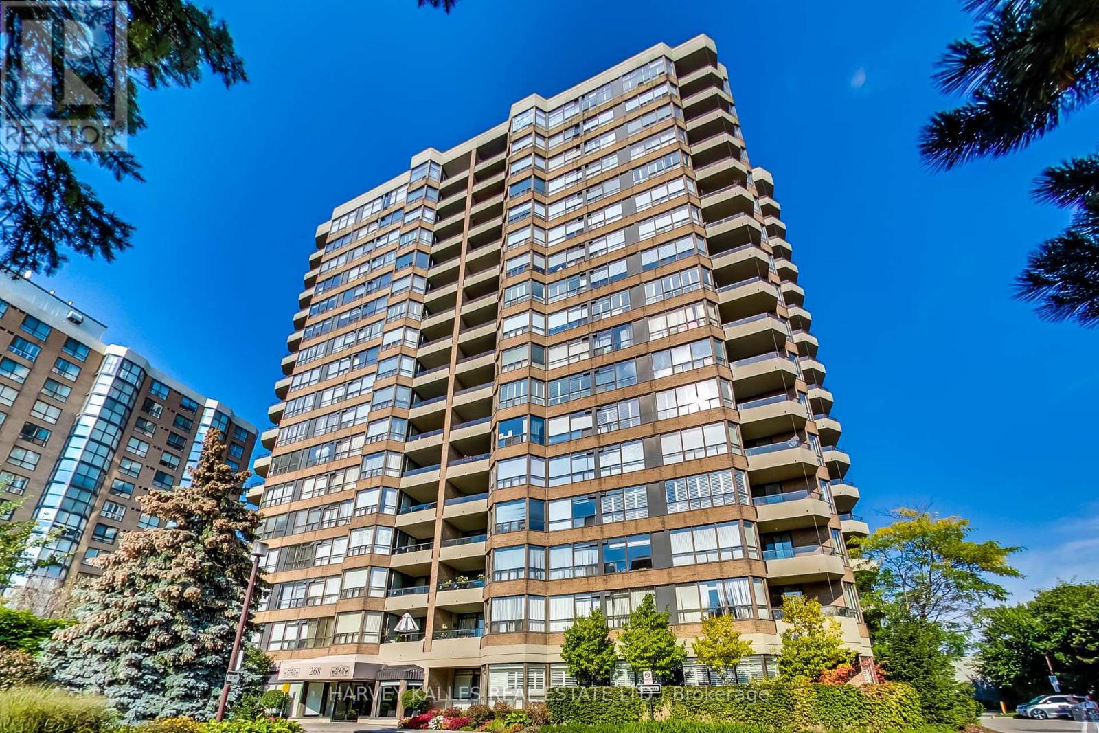 Brown-brick condominium tower with projecting balconies, framed by trees against a clear blue sky.