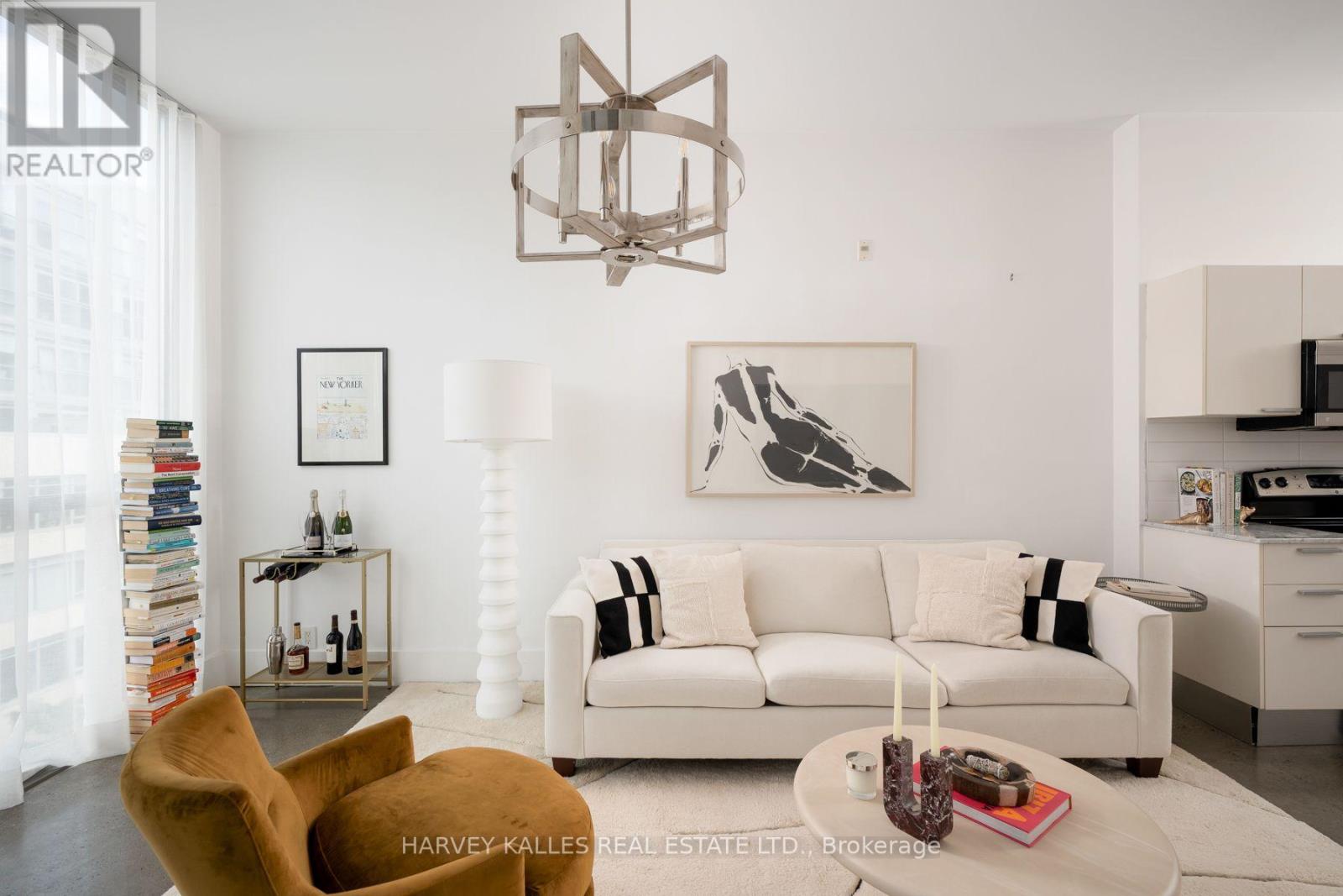 Staged living room with a white sofa, mustard lounge chair, and geometric chandelier beside an open kitchen.