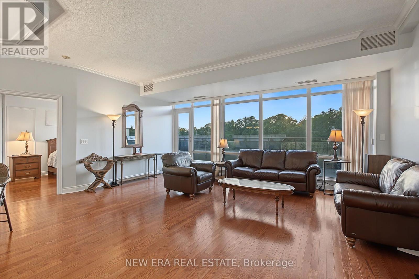 Furnished living room with hardwood floors, leather seating, and tall windows overlooking trees.