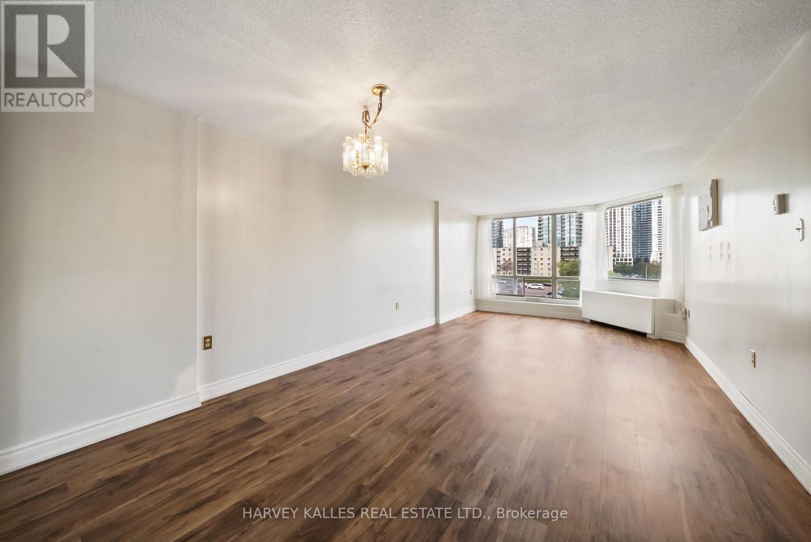 Empty apartment living room with dark wood floors, a chandelier, and large windows overlooking city buildings.