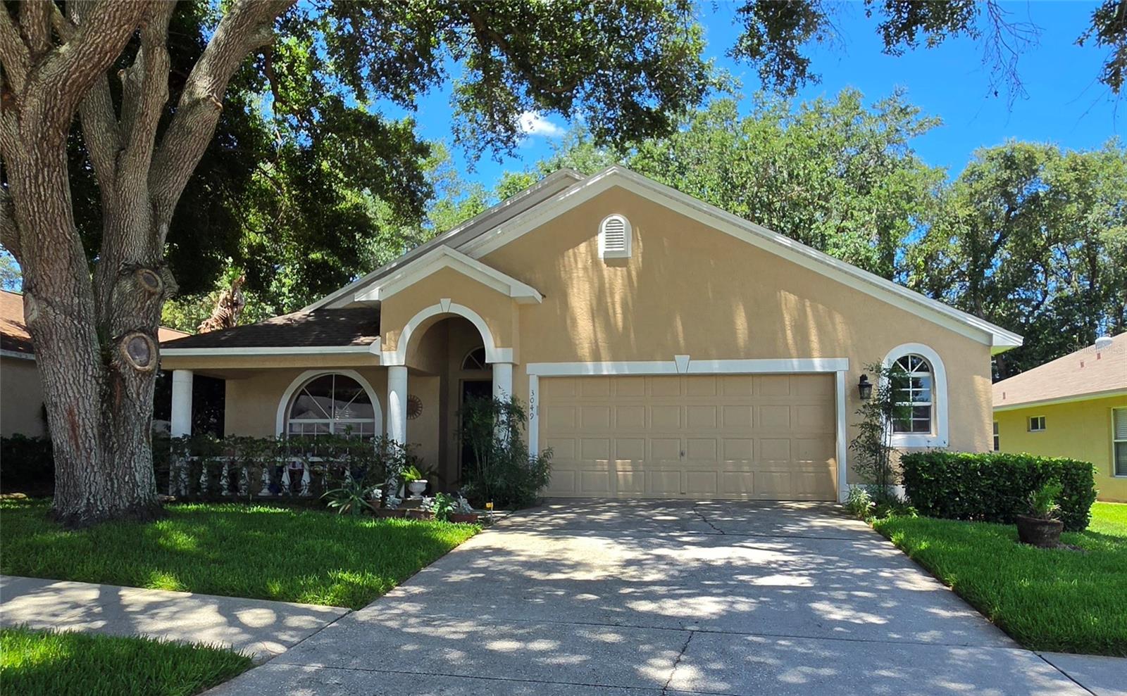 Tan single-story home with a two-car garage and covered porch beneath a large oak tree.