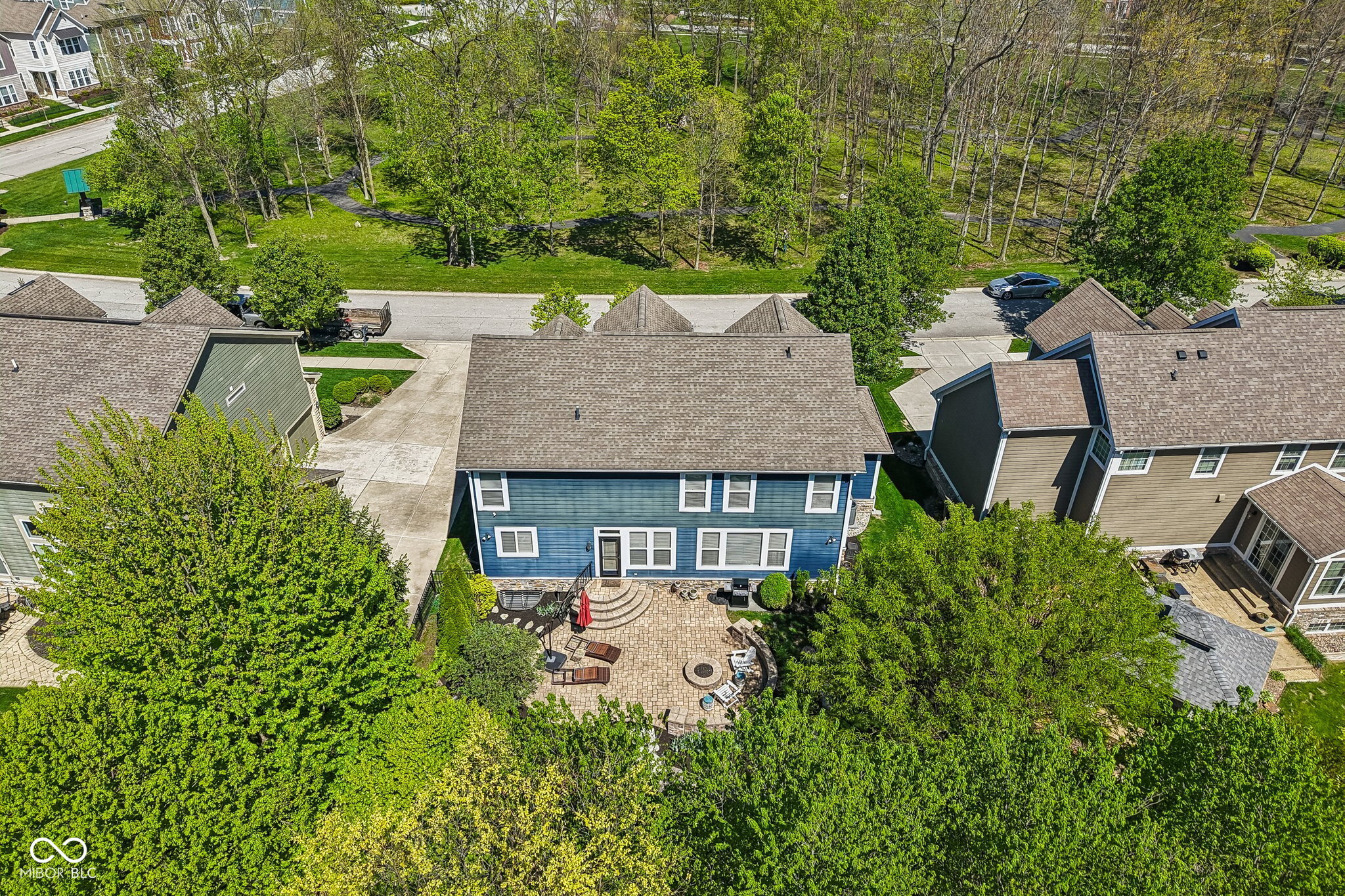 Aerial view of a blue home’s rear patio, with a fire pit and wooded walking path beyond.