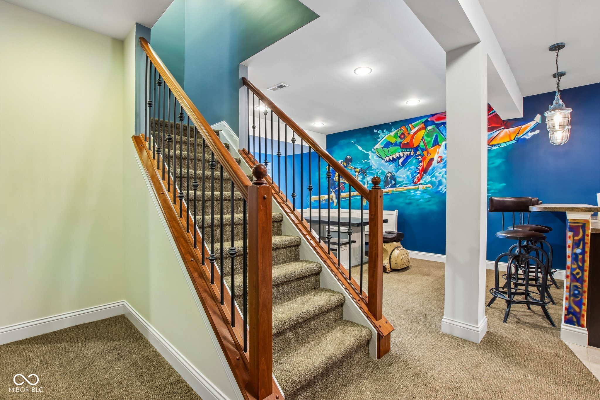Finished basement stairs with wood railings beside a game room featuring a colorful aquatic mural and bar seating.