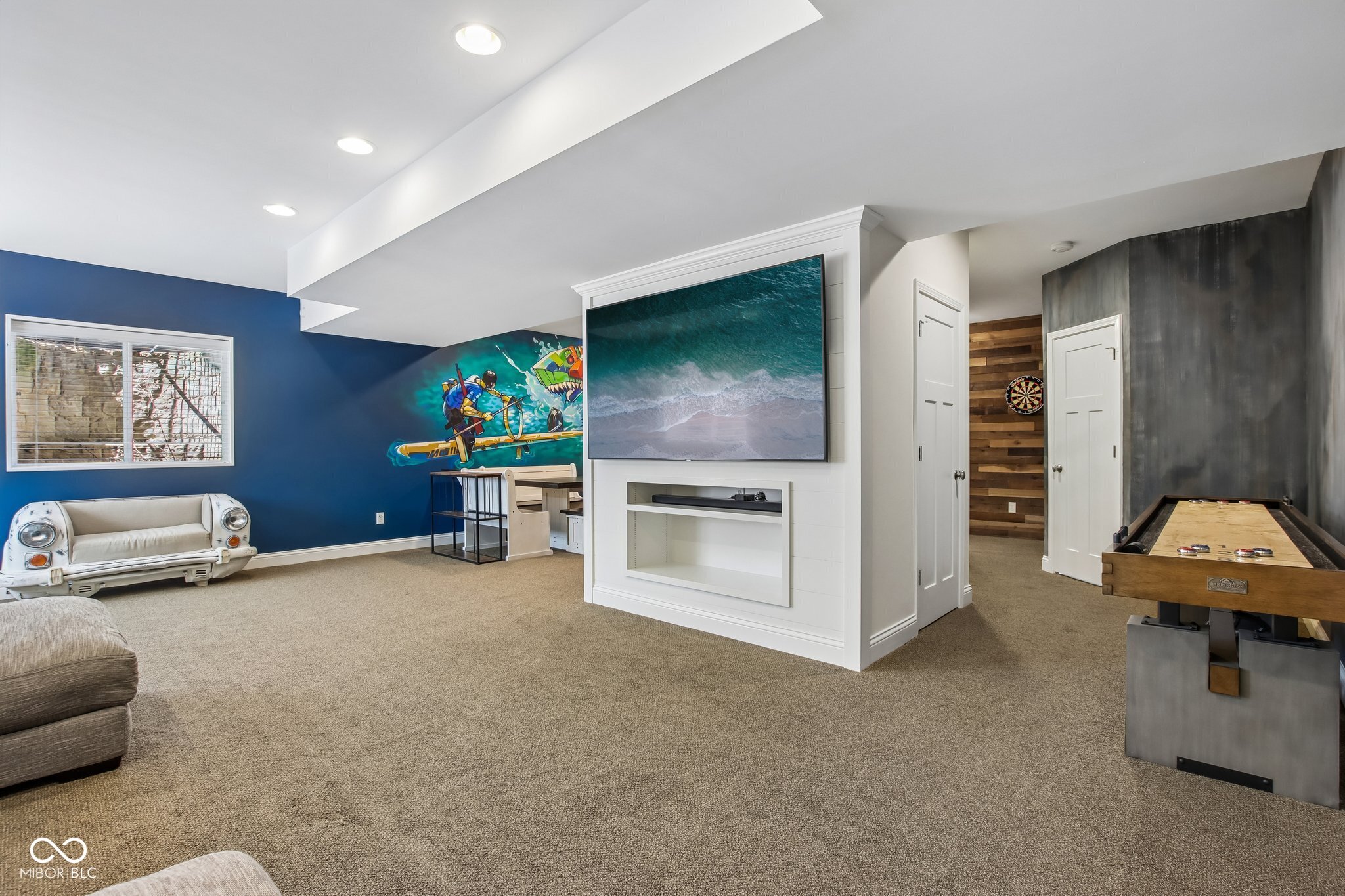 Basement recreation room with a shuffleboard table, wall-mounted TV, dartboard, and colorful mural.