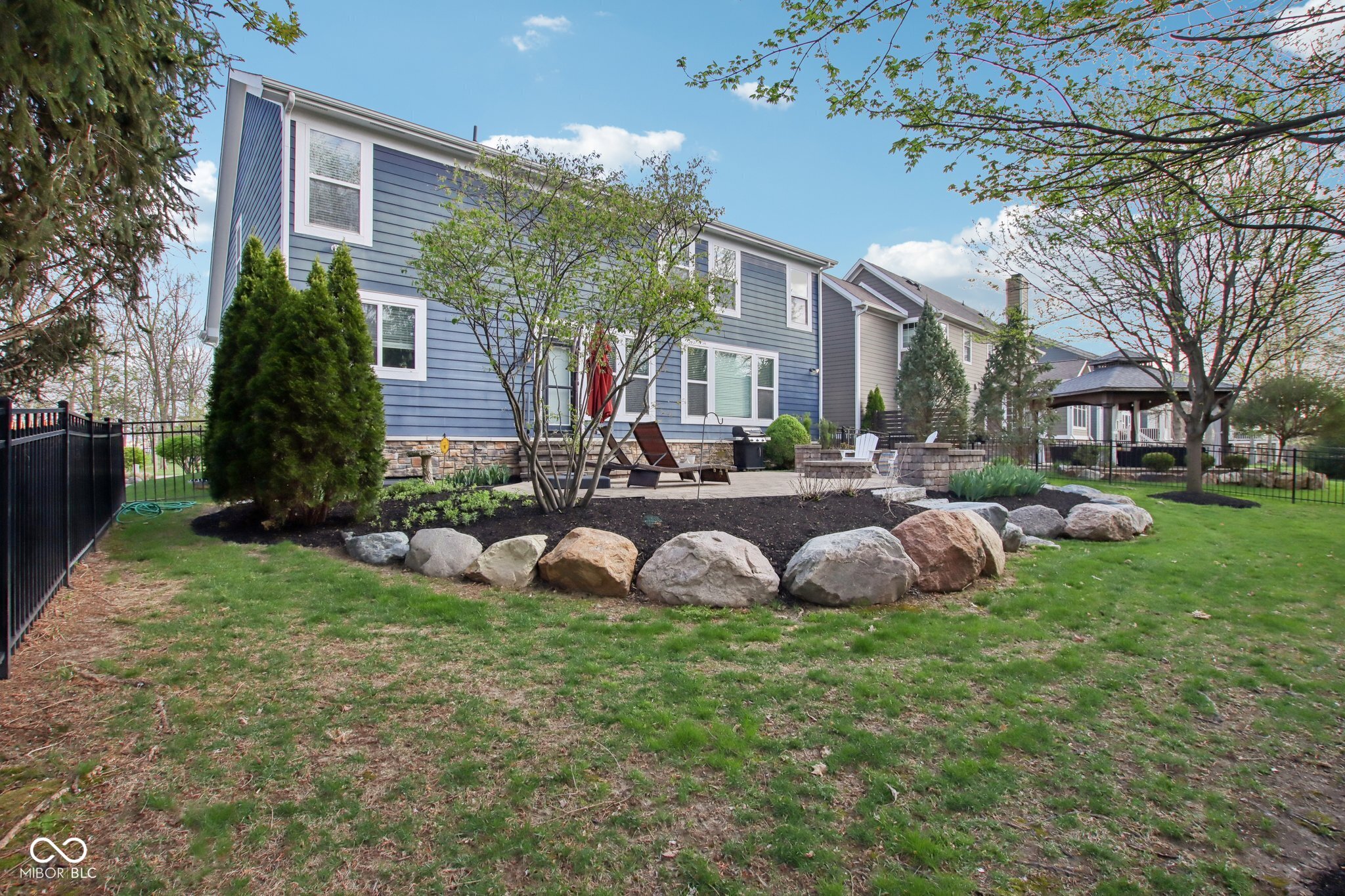 Blue two-story home with a landscaped backyard, stone-edged garden bed, patio seating, and covered gazebo.