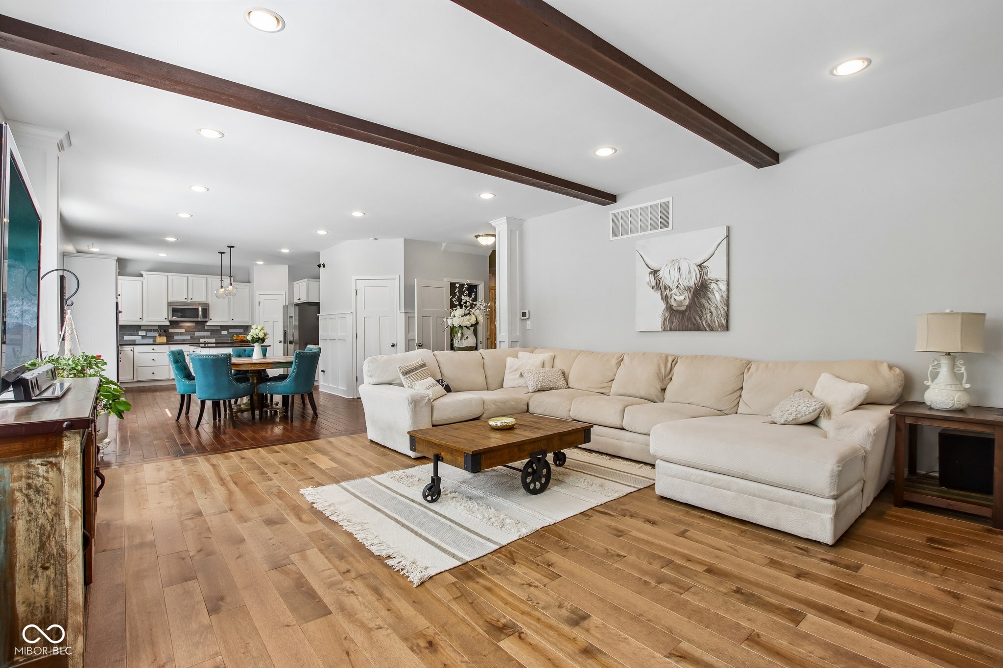 Open-plan living room with a cream sectional, dark wood beams, and kitchen dining area.