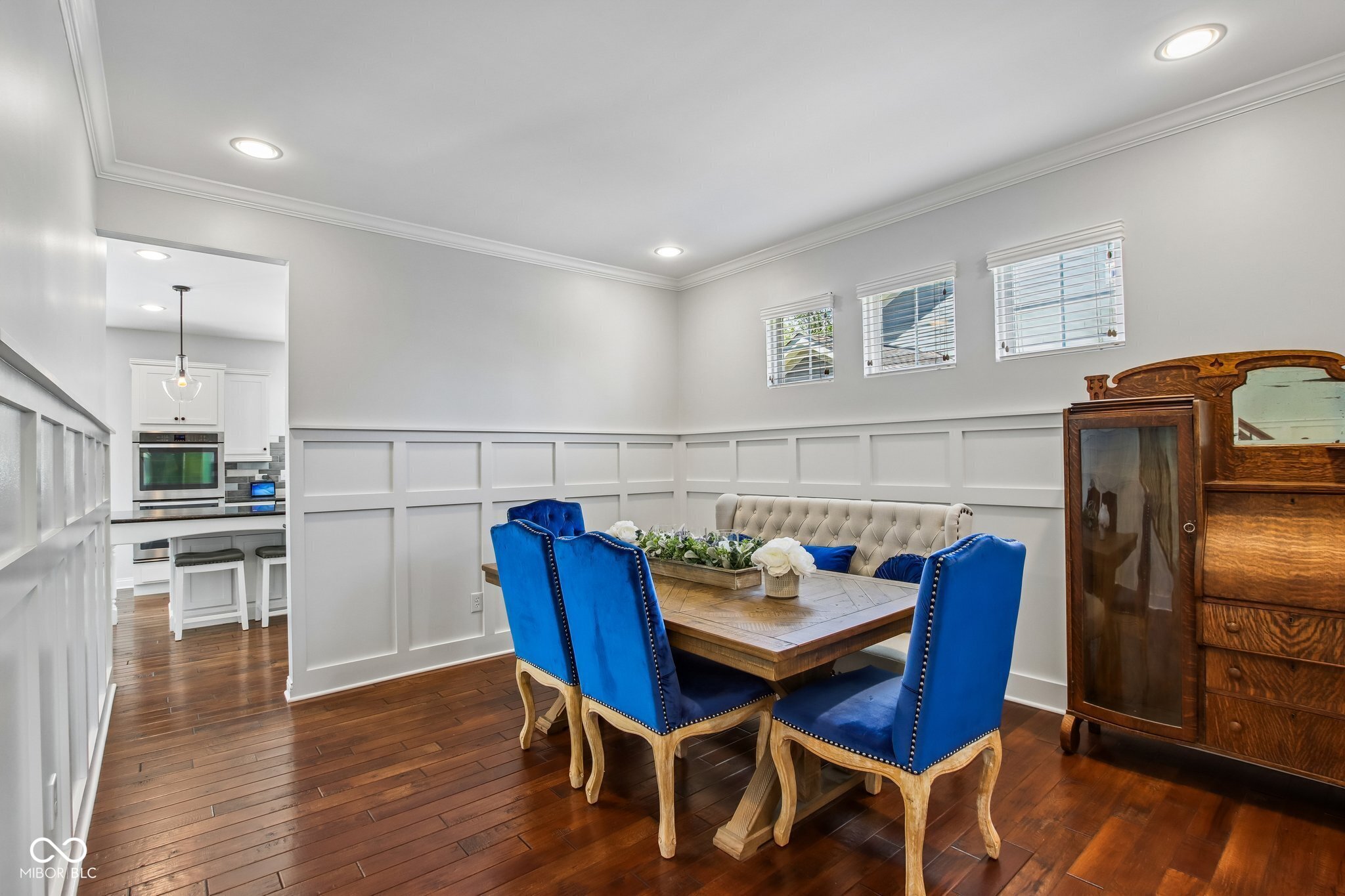 Dining room with a wooden table, bright blue chairs, white paneling, and an antique cabinet.