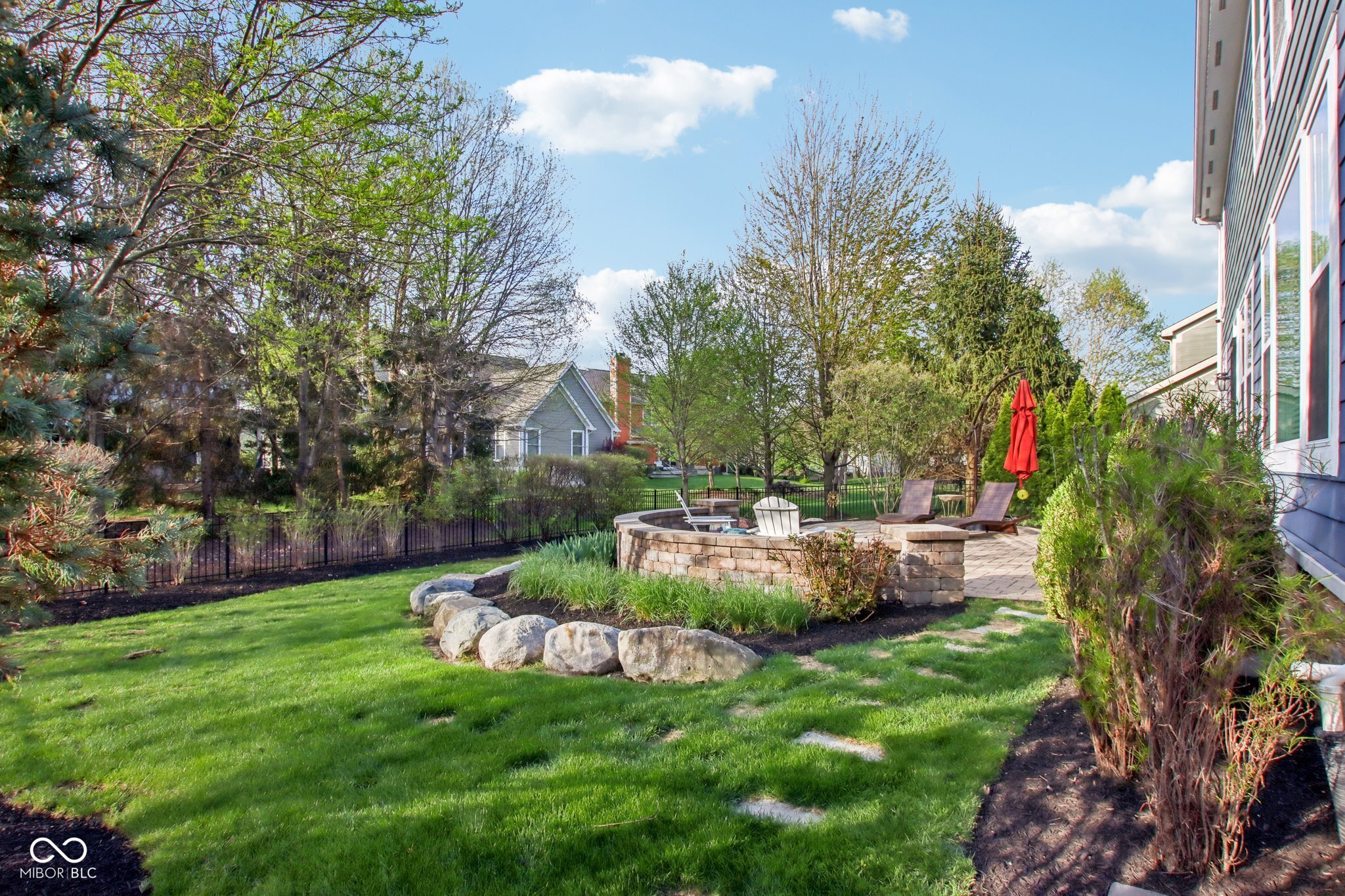Landscaped backyard with circular stone fire pit seating, a paved patio, and a closed red umbrella.