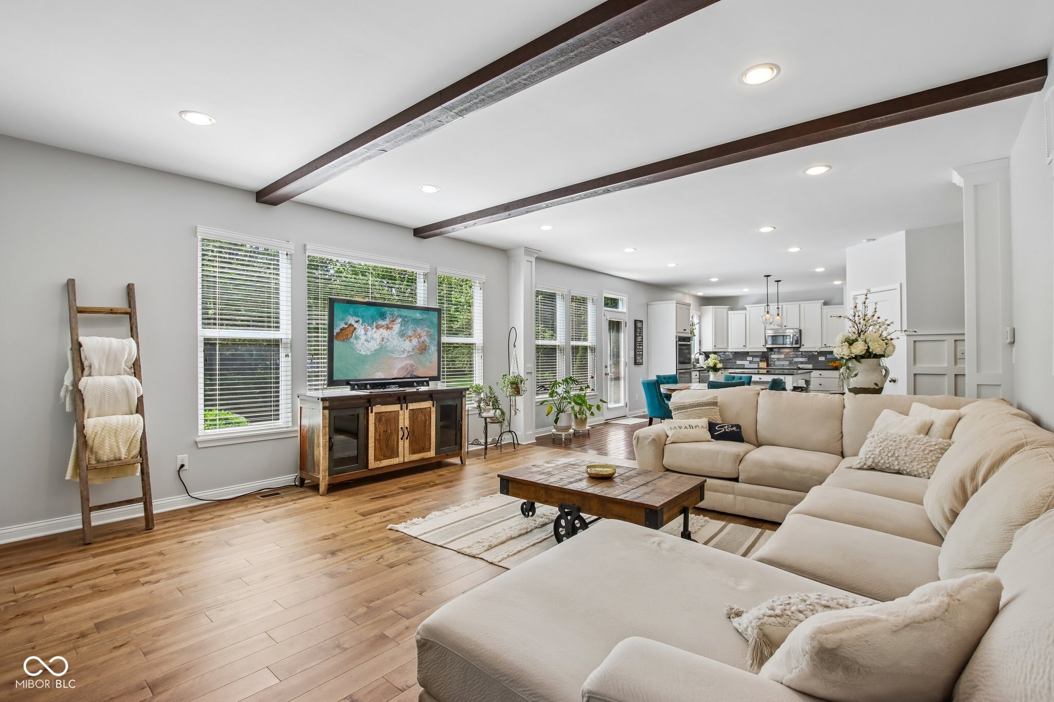 Open-plan living room with a cream sectional, wood floors, dark ceiling beams, and a white kitchen beyond.