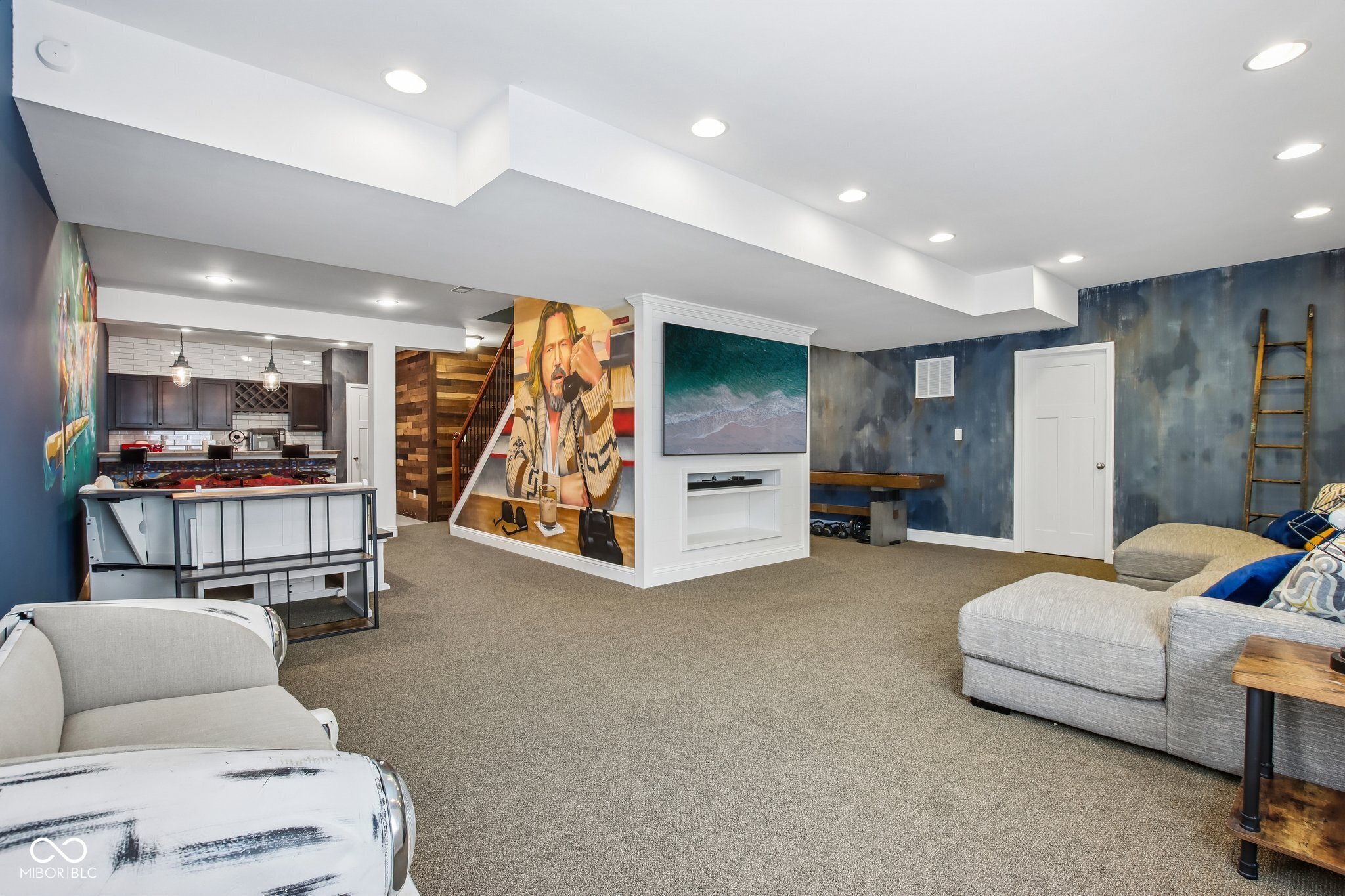 Furnished basement recreation room with gray seating, a fireplace, kitchenette, and large portrait mural.
