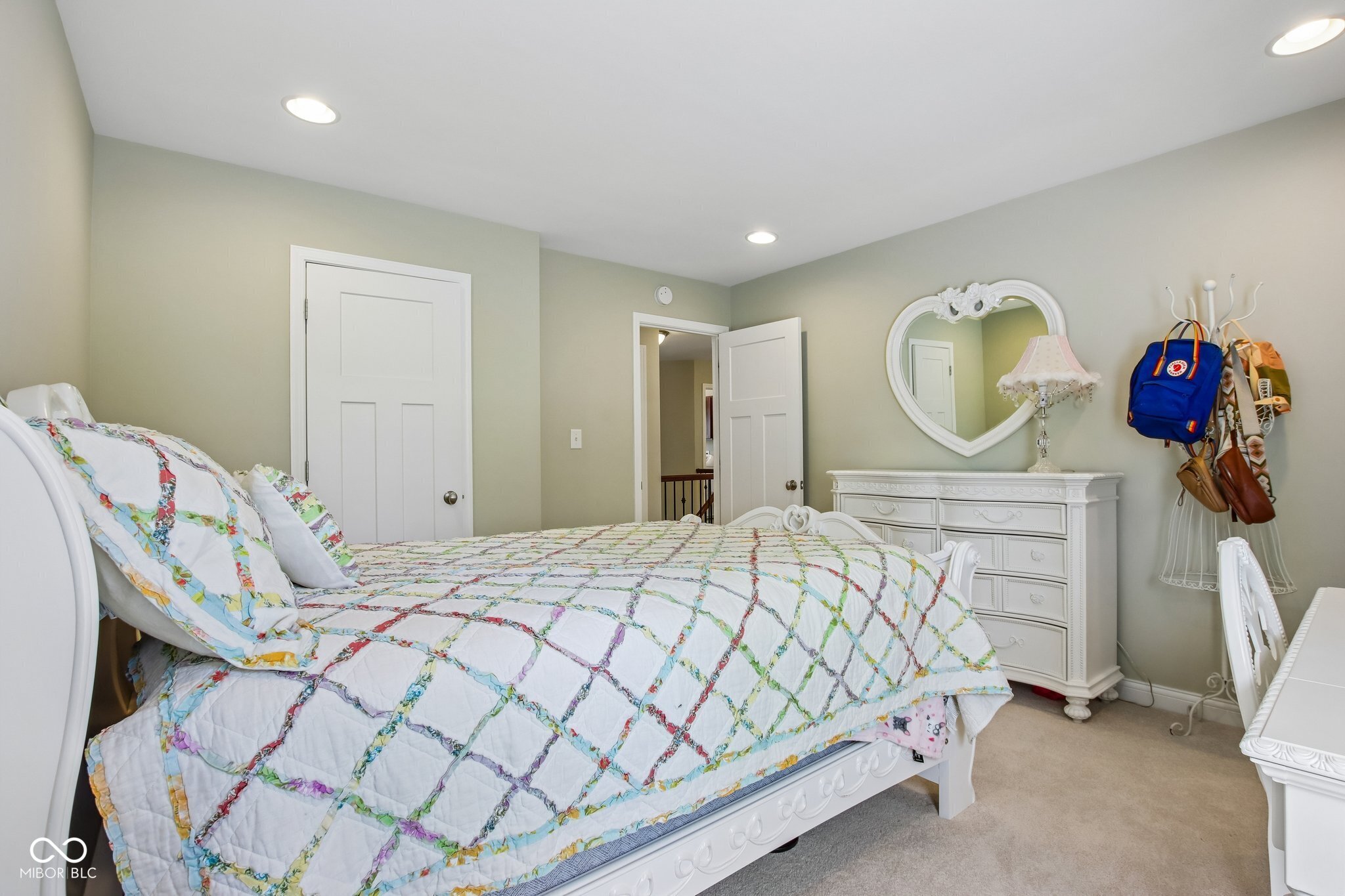Bedroom with a colorful patchwork quilt, heart-shaped mirror, and white dresser.