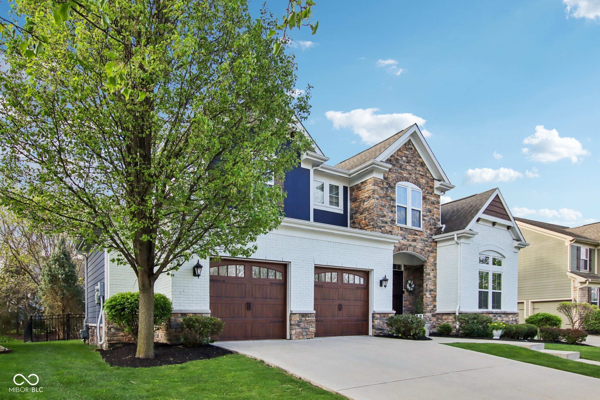 Two-story house with white brick, stone accents, double garage, and a leafy tree in front.