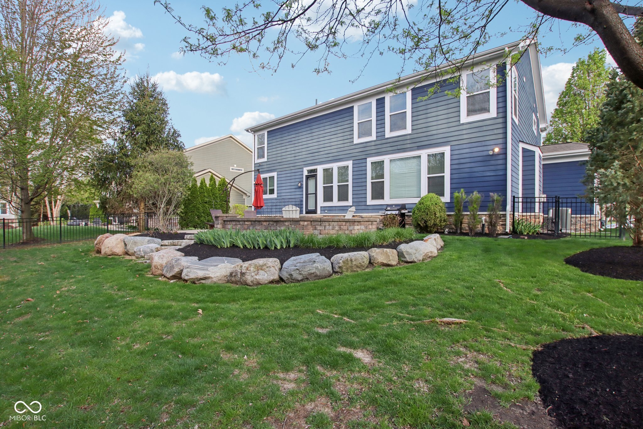 Blue two-story home behind a spacious lawn, with a raised stone patio and curved rock-edged garden bed.