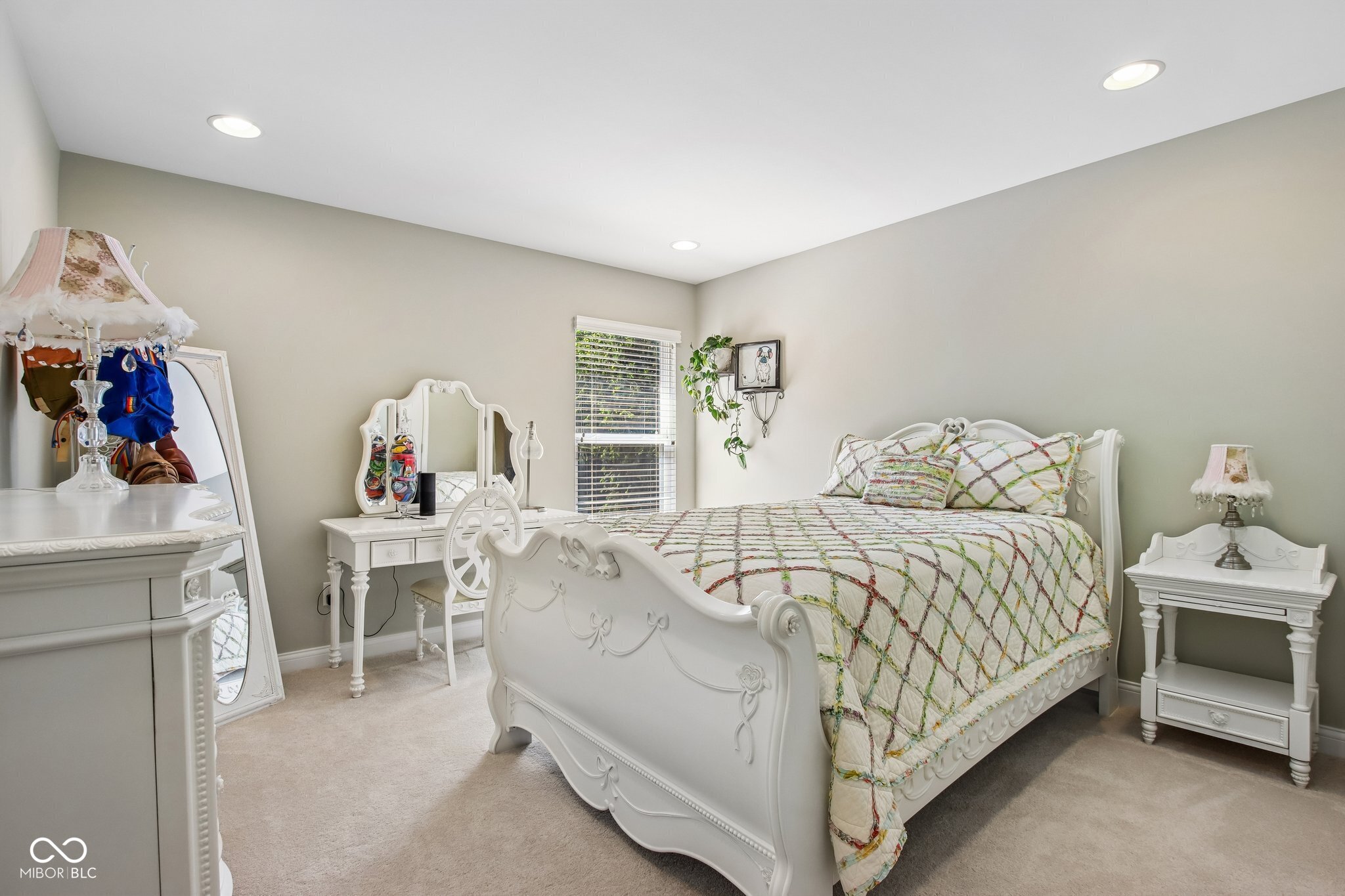 Bedroom with an ornate white bed, multicolored quilt, matching vanity, and window with blinds.