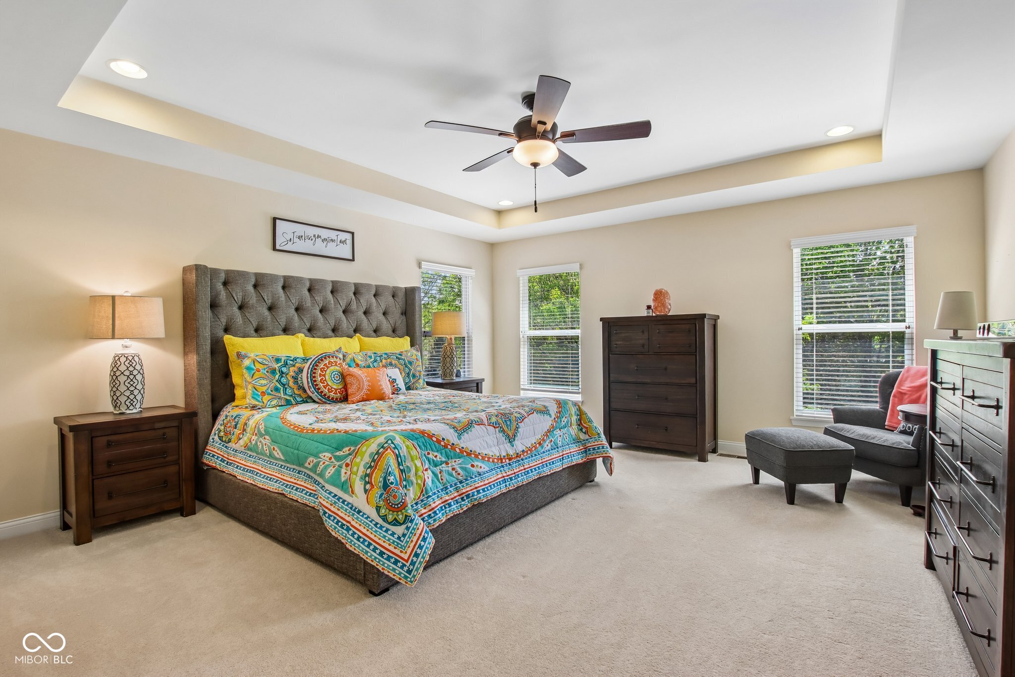Furnished bedroom with a colorful patterned bedspread, tufted headboard, tray ceiling, and windows overlooking greenery.